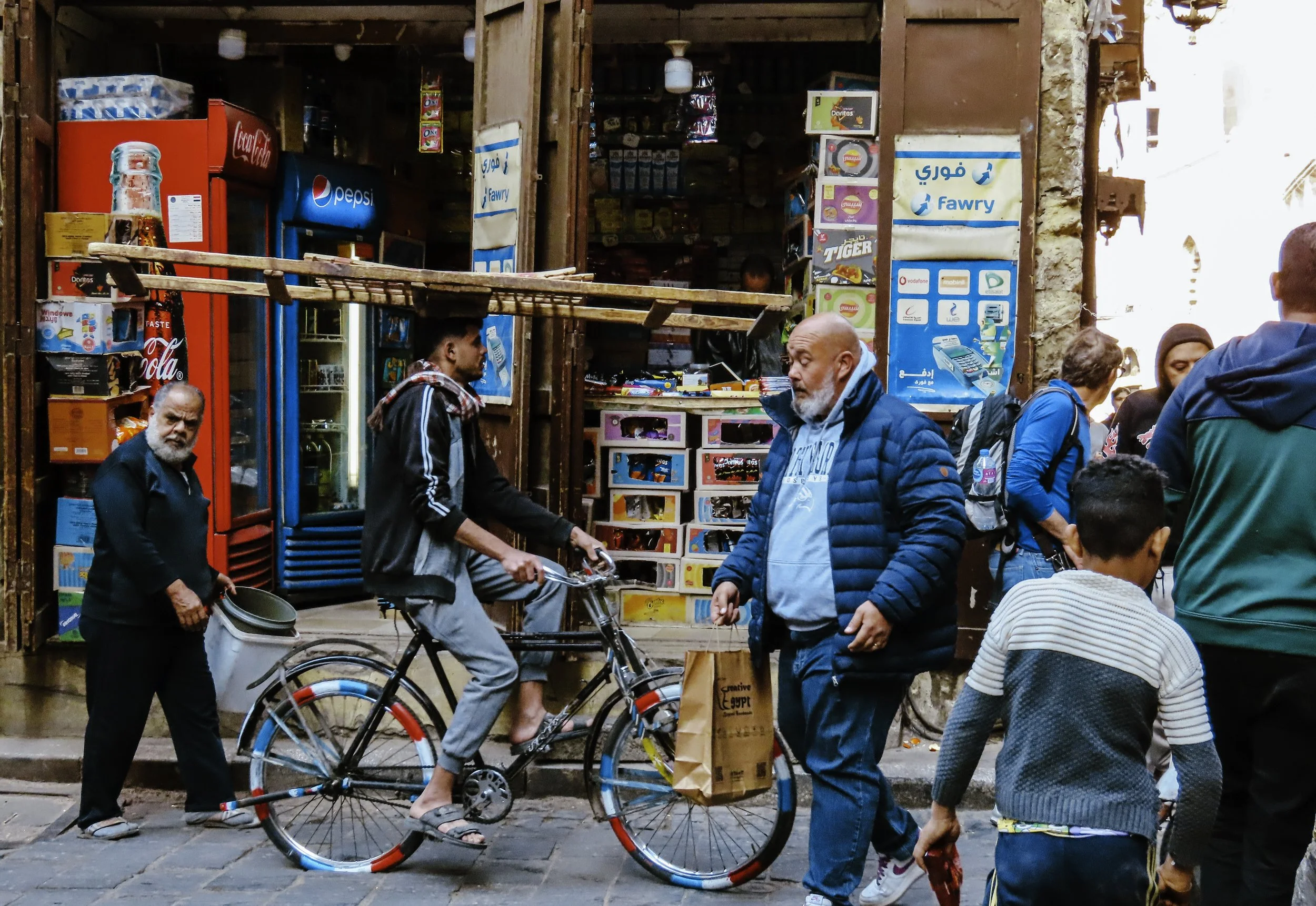 Scene di strada con persone che camminano e un ragazzo che pedala su una bicicletta, negozio con bibite in secondo piano.