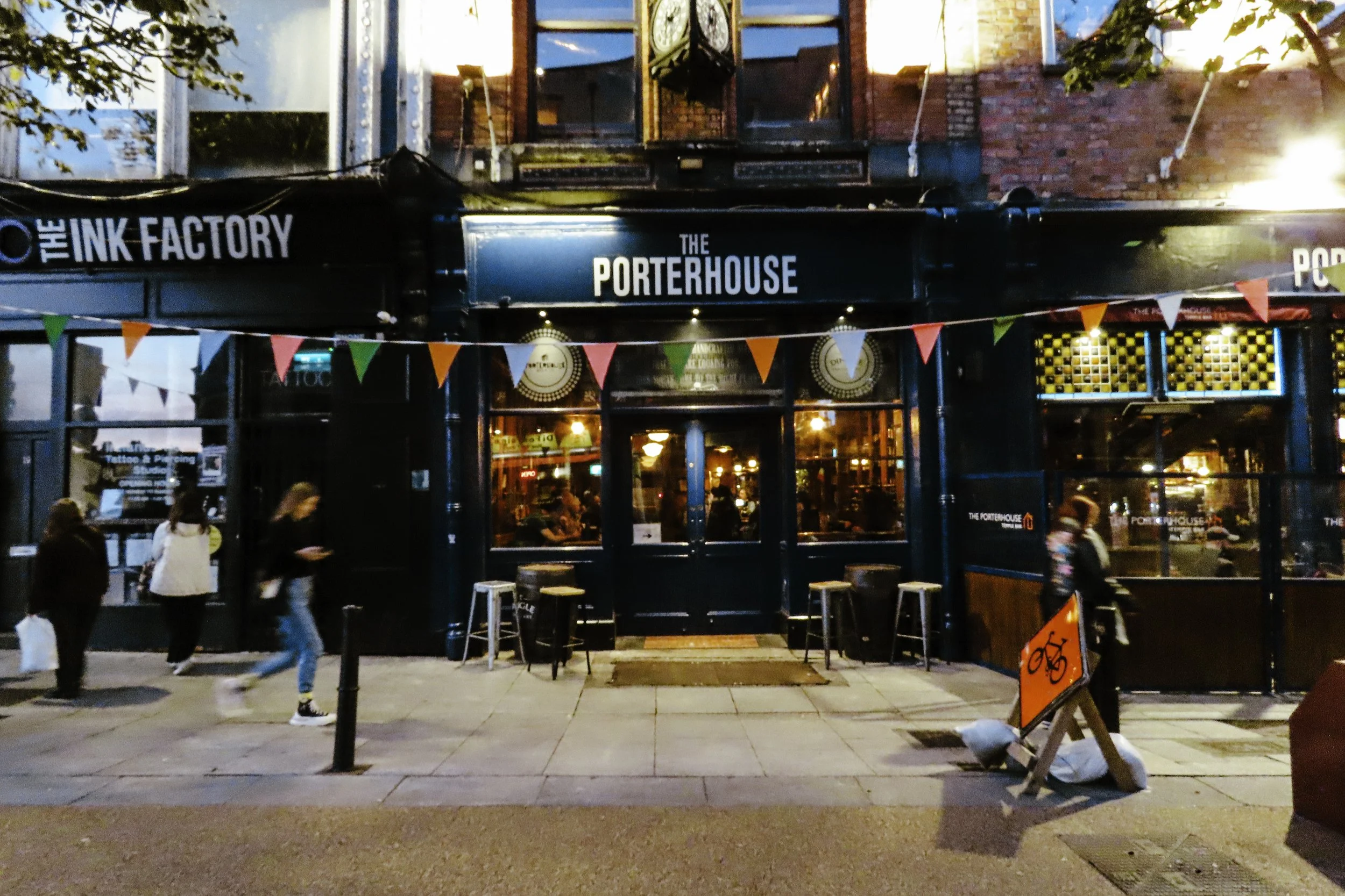 Facciata di un pub di sera chiamato 'The Porterhouse'. Sul cartellone ci sono decorazioni di bandierine di vari colori e alcune persone passeggiano davanti all'ingresso, che è tra due grandi vetrate illuminate. C'è anche un cartello con simbolo di bi