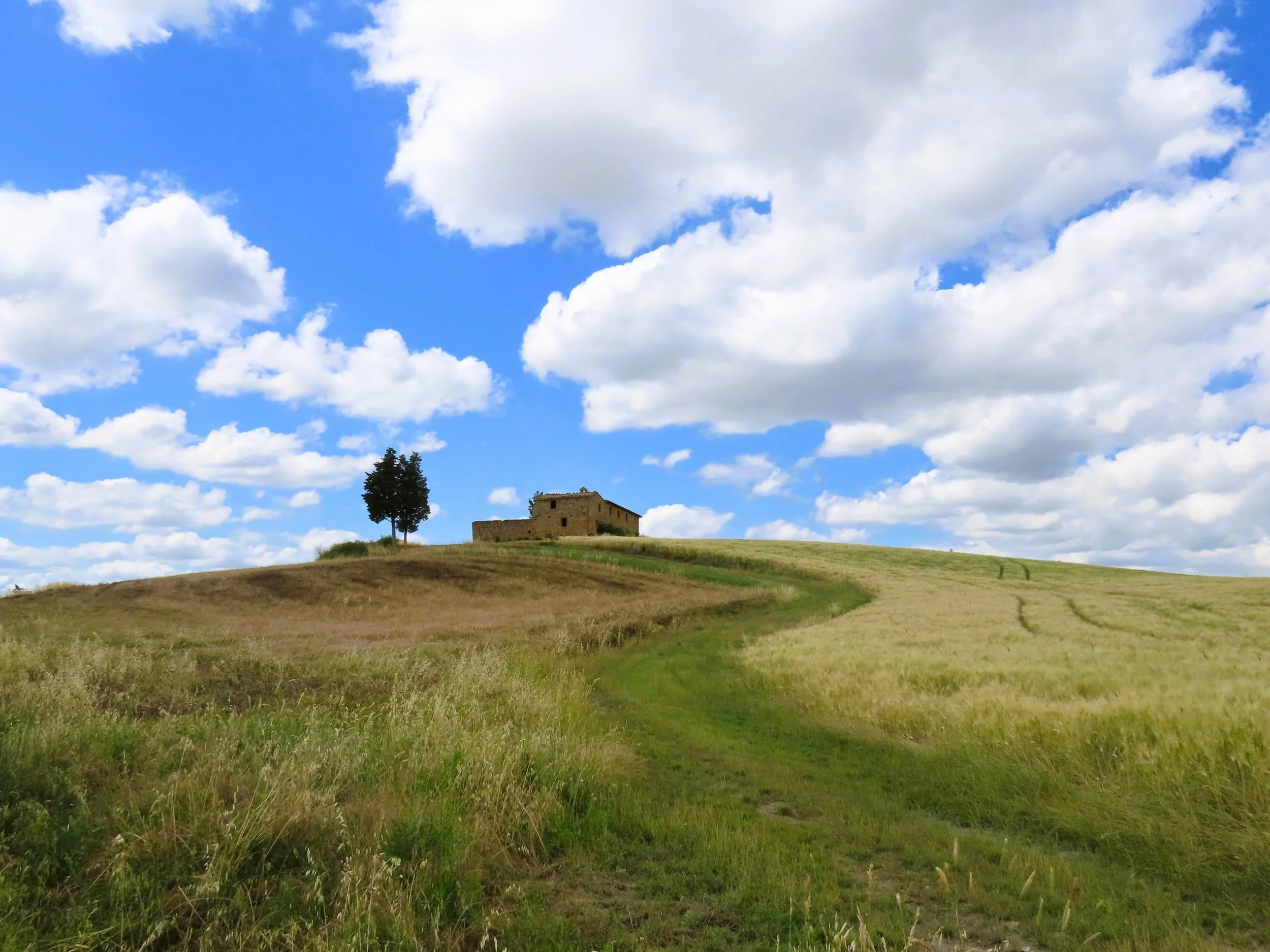 Paesaggio rurale con collina, casa in pietra e un albero, sotto un cielo blu con nuvole