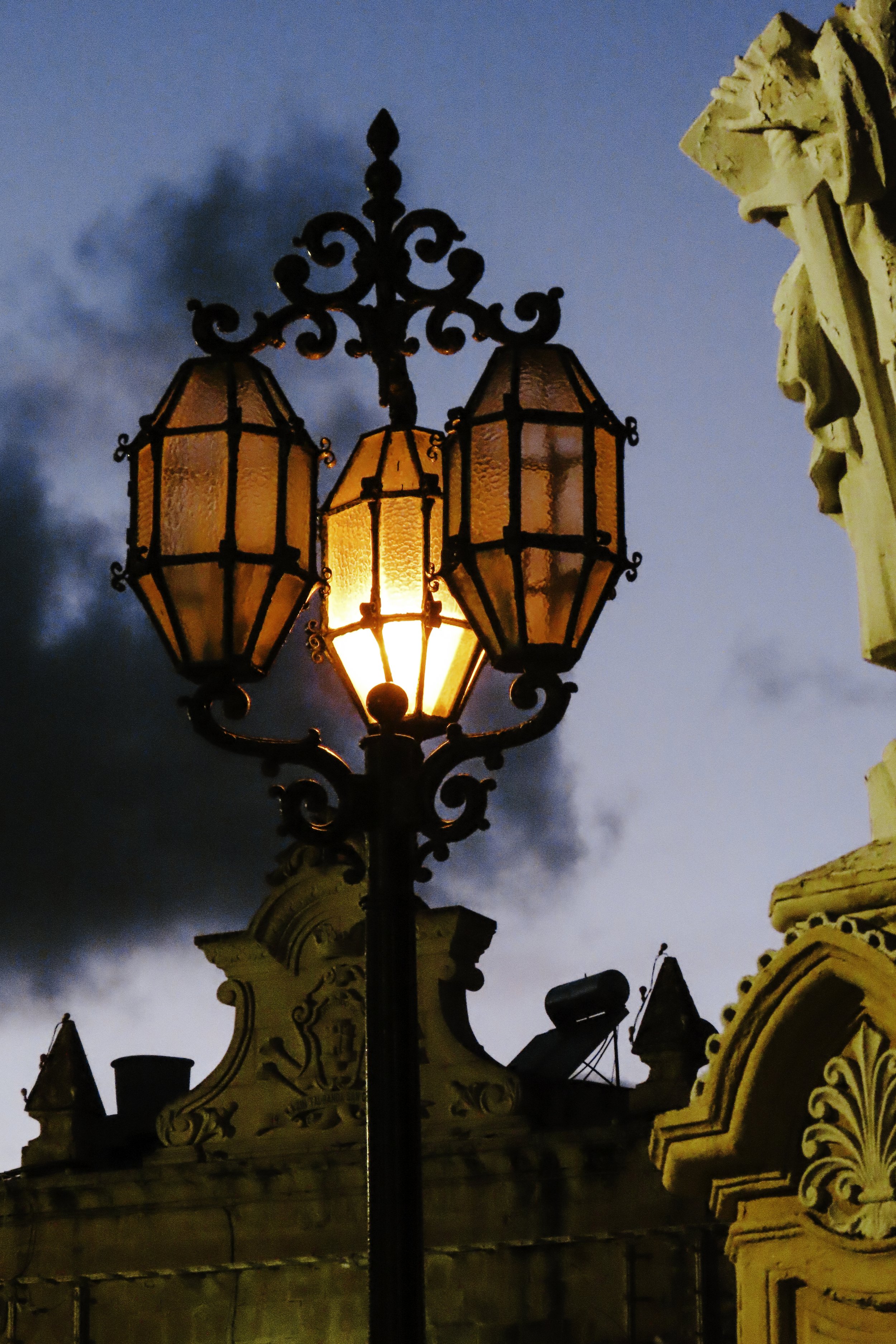 Lampione in stile antico con tre luci accese, al tramonto, contro un cielo nuvoloso.