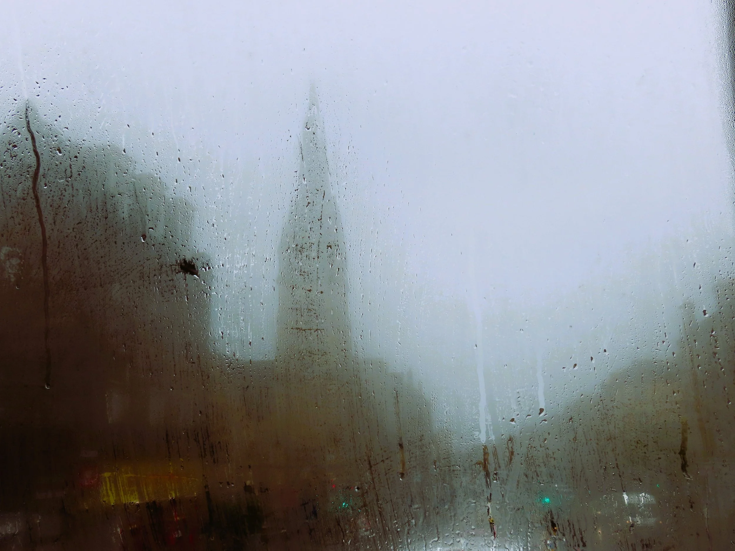 Finestra appannata con gocce di pioggia che oscurano una vista urbana con edifici e un traliccio in lontananza.