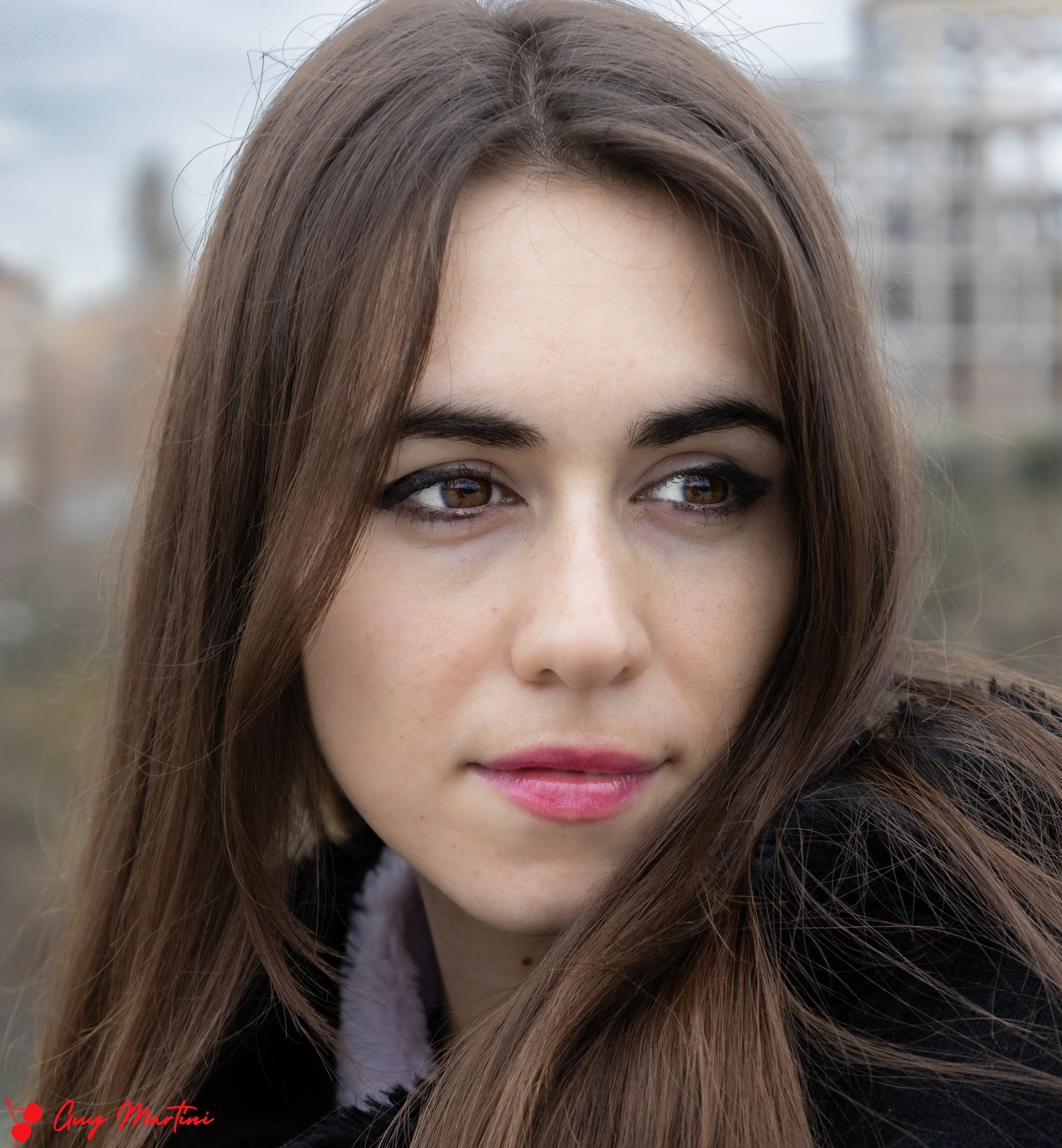Primo piano di una giovane donna con capelli castani, occhi marroni, trucco leggero e sorriso leggero, con sfondo sfocato di edifici urbani.
