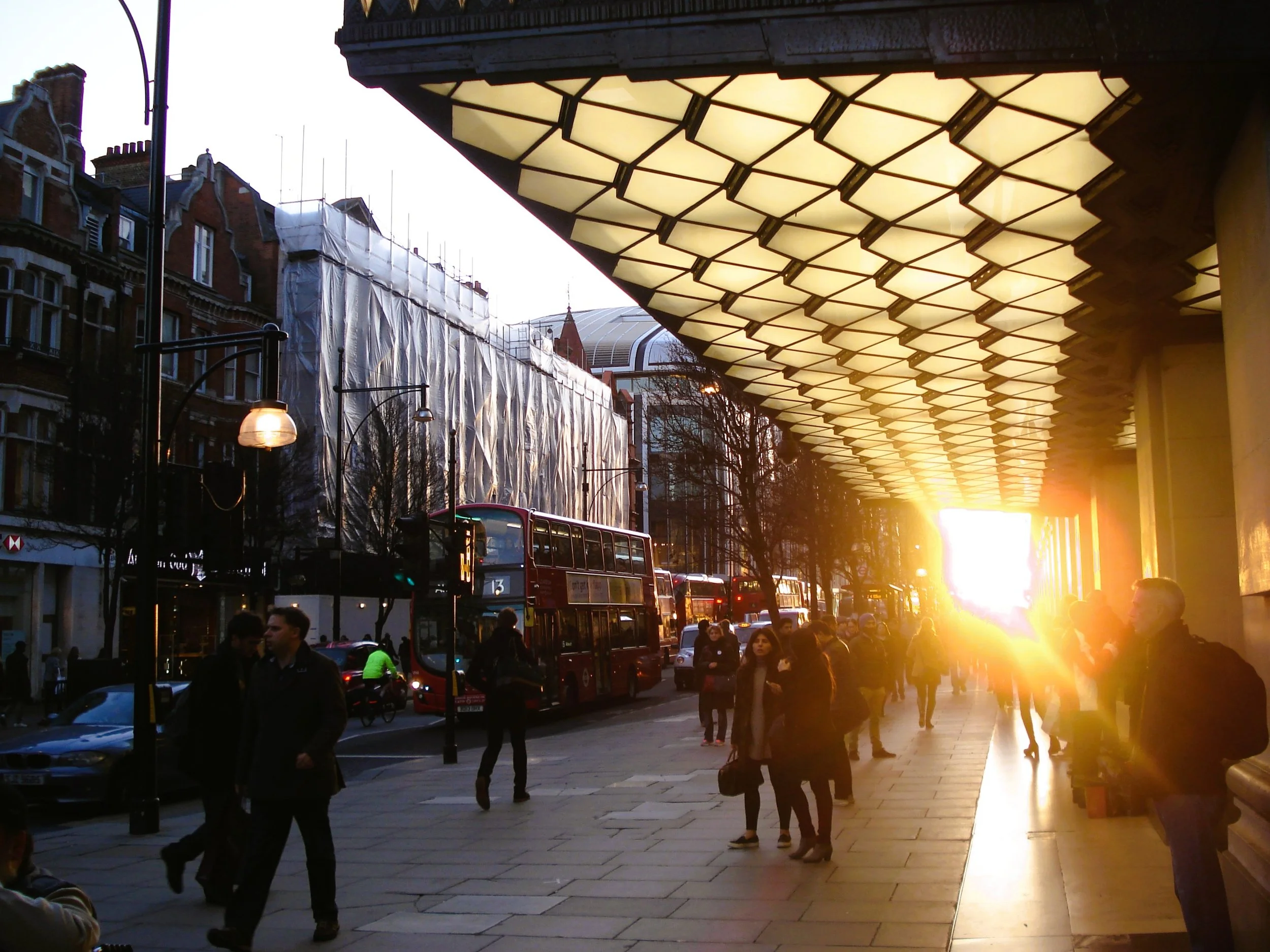 Immagine di una strada cittadina al tramonto, con molte persone che camminano e alcuni bus rossi. Sotto una tettoia luminosa si vede il sole al tramonto.