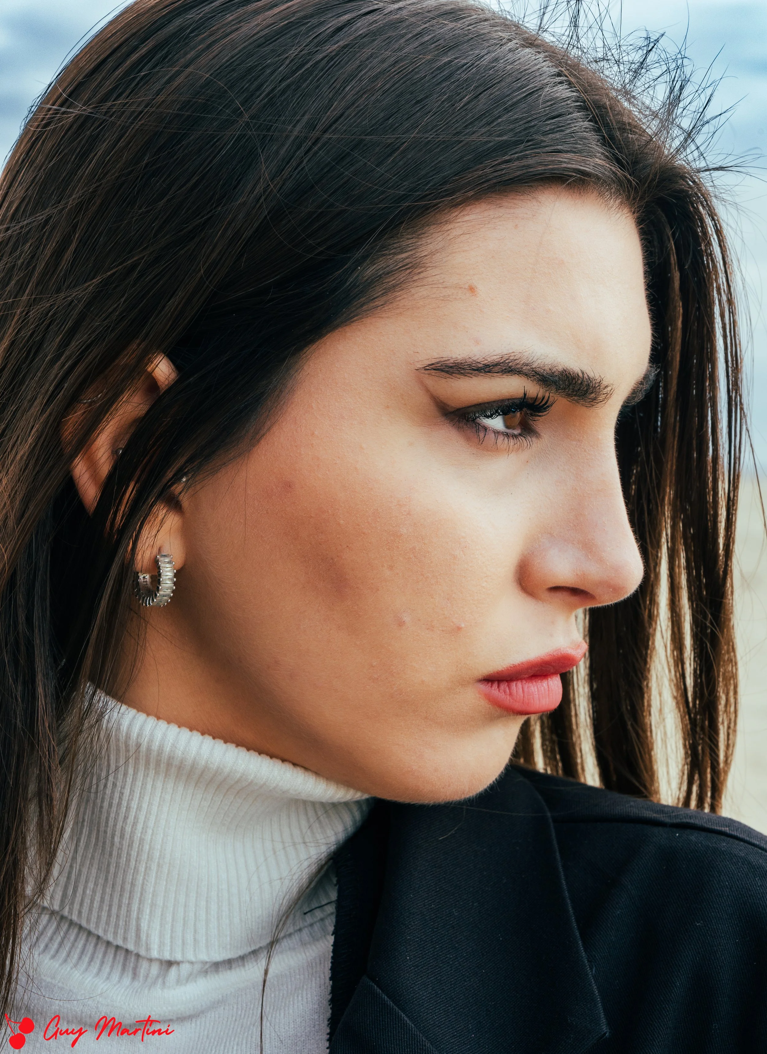 Ritratto di una donna con capelli marroni, pelle chiara, indossa orecchini d'argento, maglia bianca a collo alto e giacca nera, con sfondo di cielo e mare.