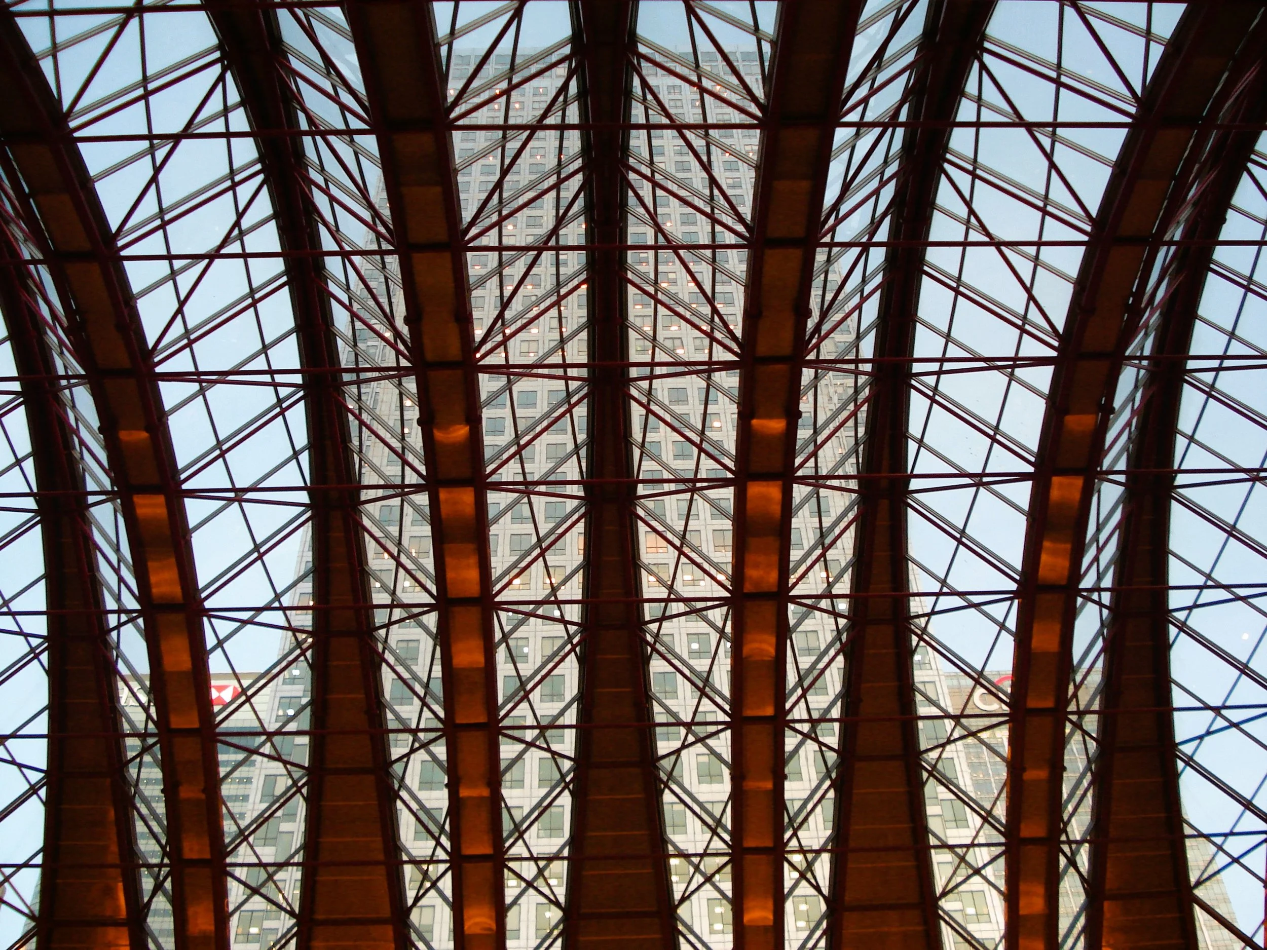 Interno di una struttura con un soffitto di vetro e ferro, con un edificio alto in vetro visibile attraverso il soffitto.