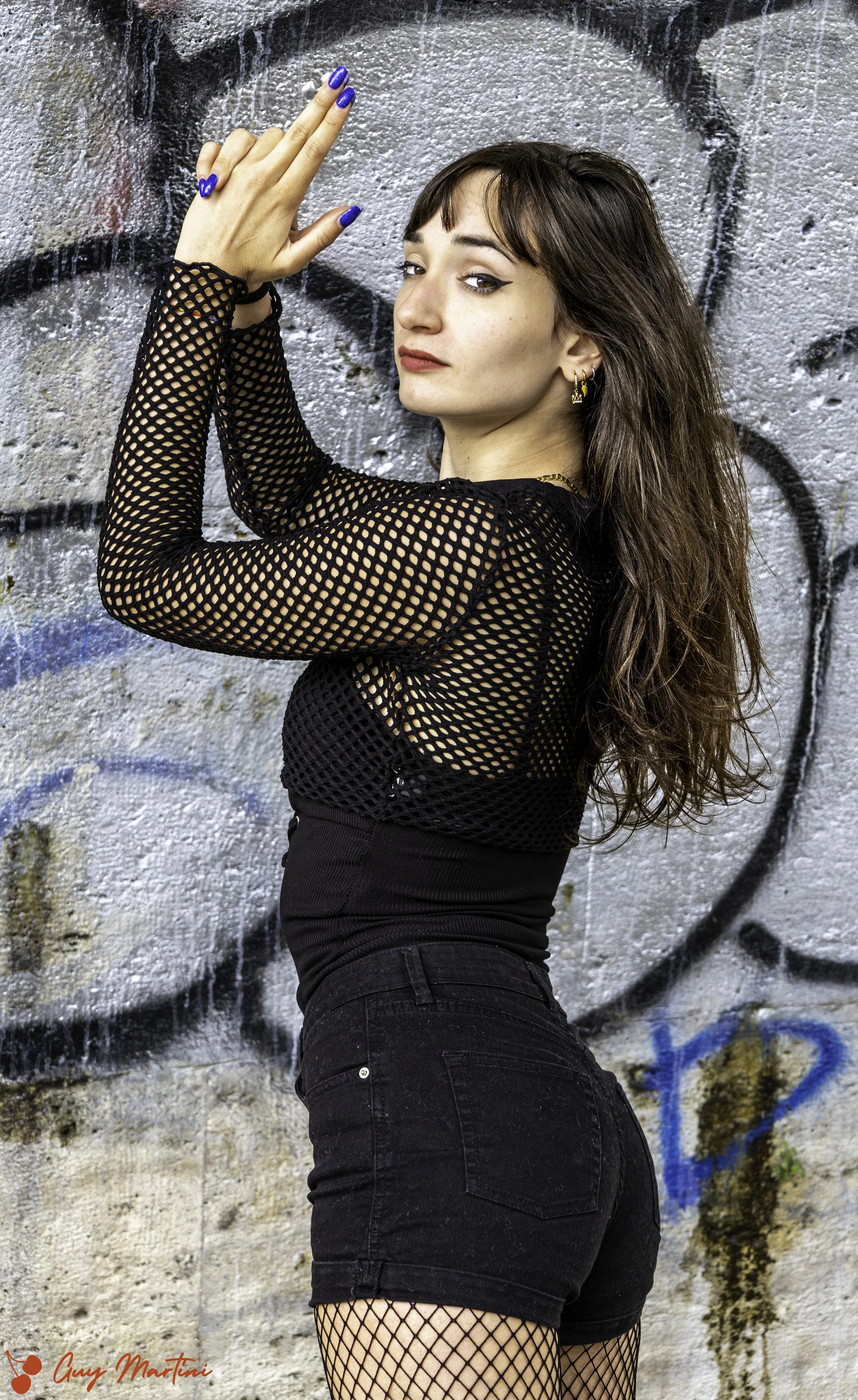 Ragazza con capelli lunghi, indossa maglietta di rete nera e pantaloncini neri, posa con braccia sollevate contro un muro con graffiti.