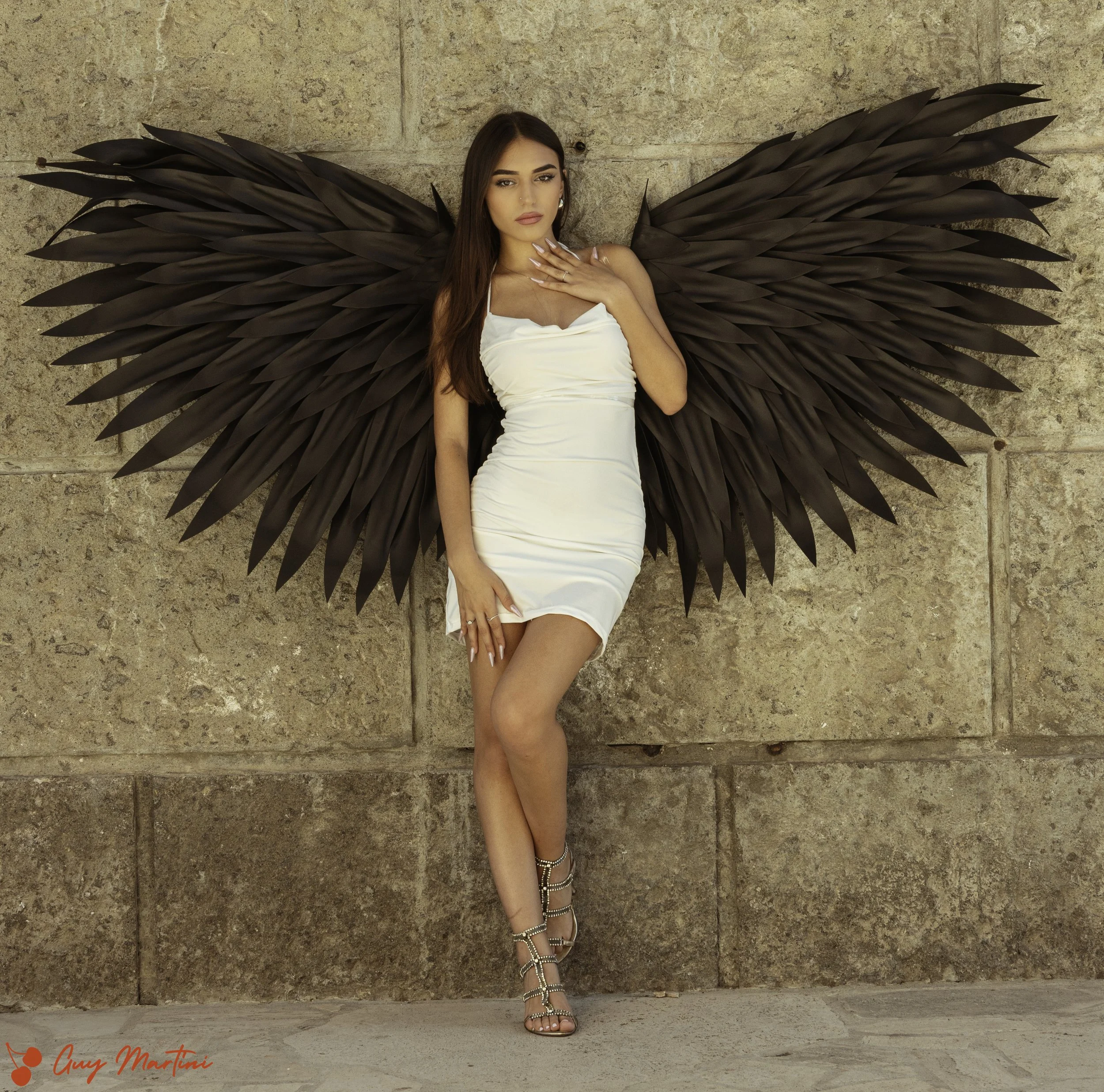 Una donna con grandi ali di piume nere, indossa un vestito bianco attillato e scarpe con cinturini, posando contro un muro di pietra.