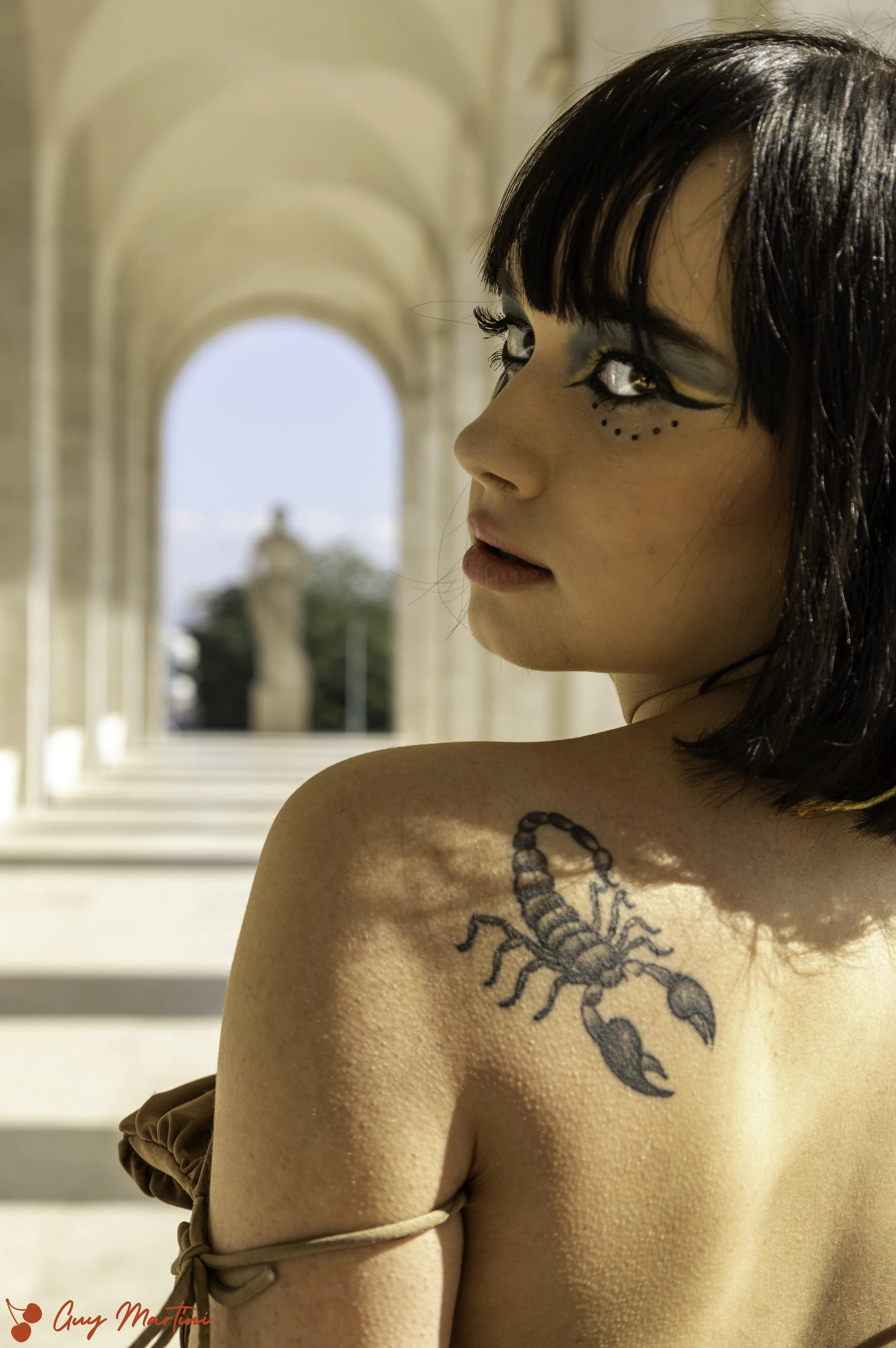 Ragazza con trucco teatrale, tatuaggio scorpione sulla spalla, ambientazione con archi di pietra.