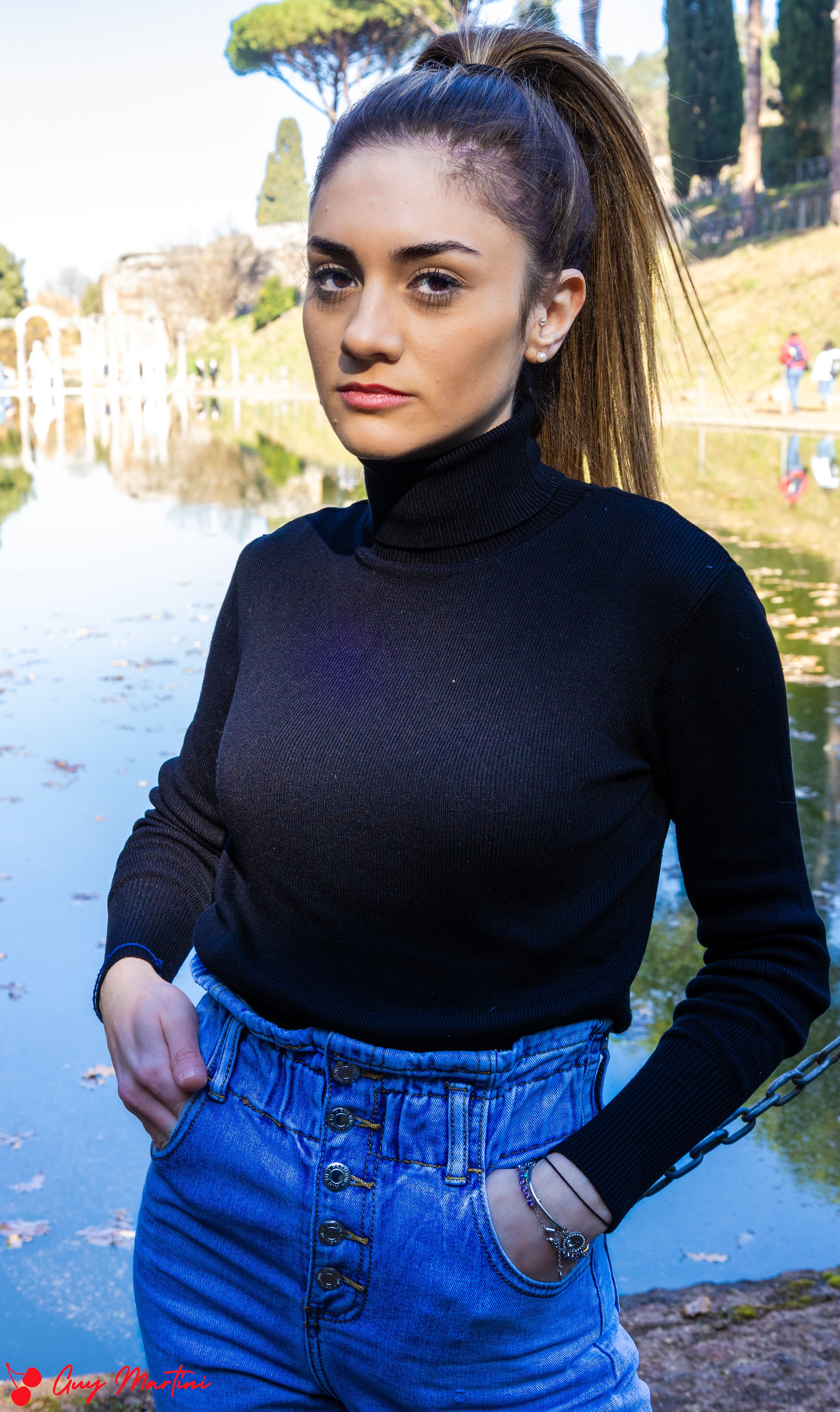 Ragazza con acconciatura alta, maglione nero, jeans a vita alta, in piedi vicino a un laghetto con alberi e persone sullo sfondo.