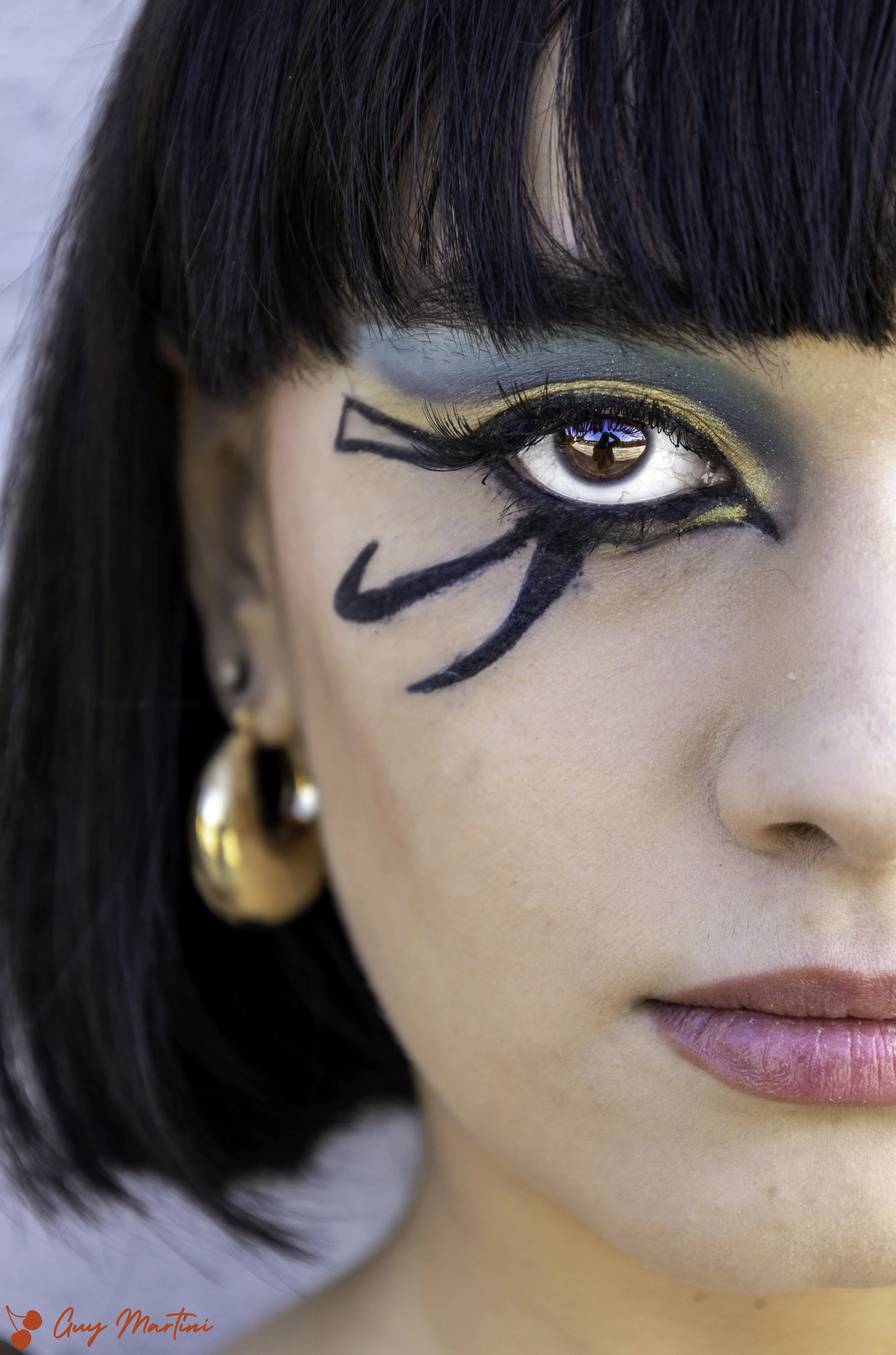 Close-up di un volto femminile con trucco artistico sugli occhi, occhi marroni, labbra leggere, orecchino d'oro, capelli corti e scuri, riflesso di una scena nel riflesso dell'occhio.