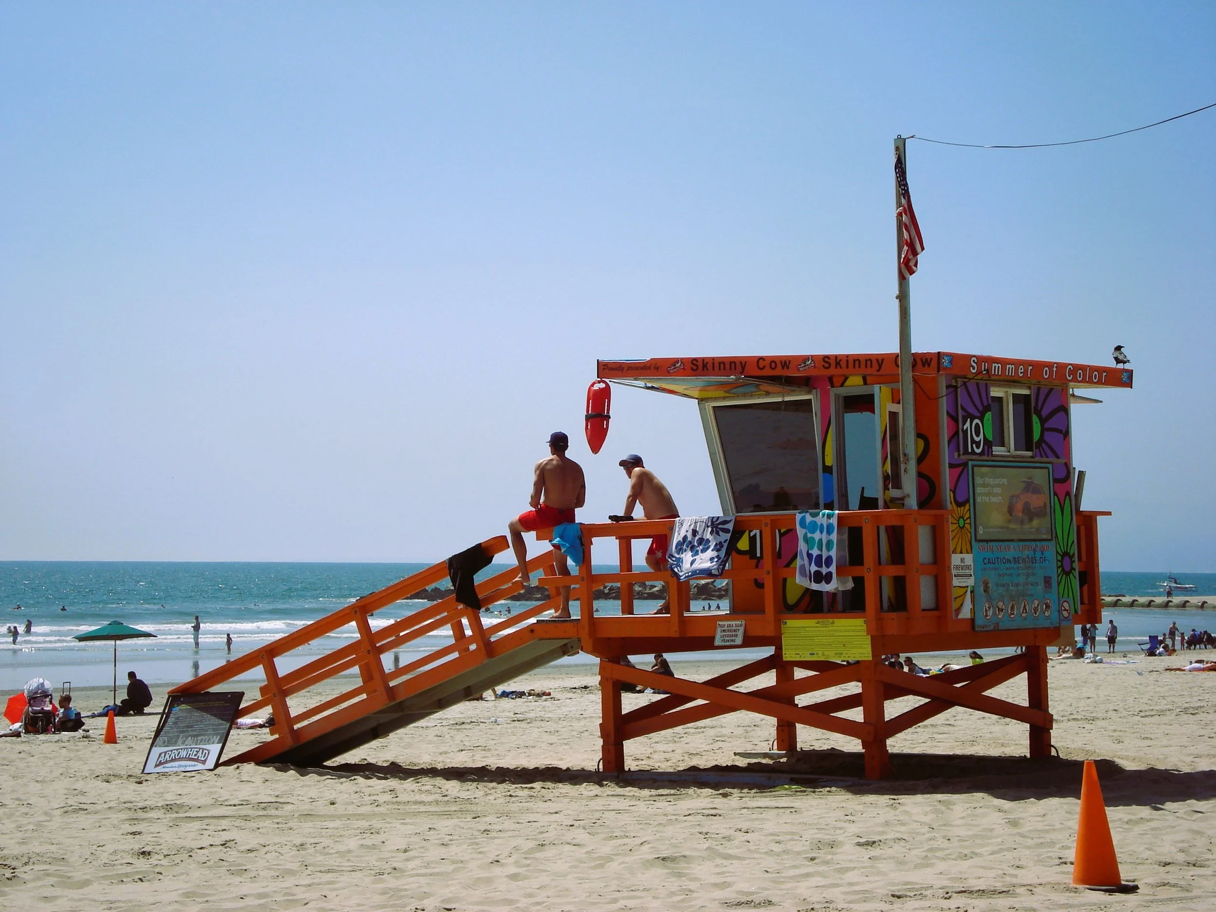 Torri di salvataggio arancioni sulla spiaggia con due bagnanti seduti sopra e persone sulla spiaggia e nel mare in background