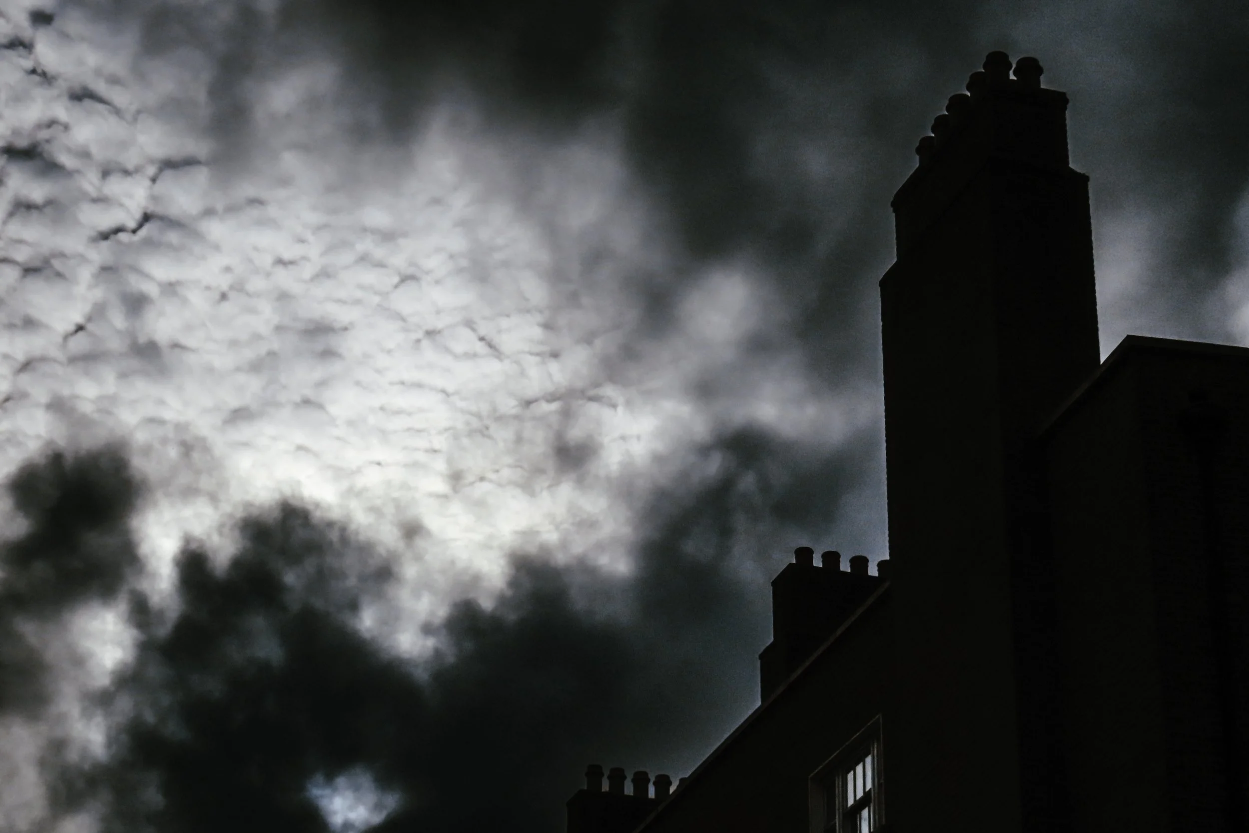 Cielo nuvoloso con un edificio in ombra, sulla destra, e un camino che si eleva verso il cielo con nuvole dense e illuminate dalla luce diffusa.