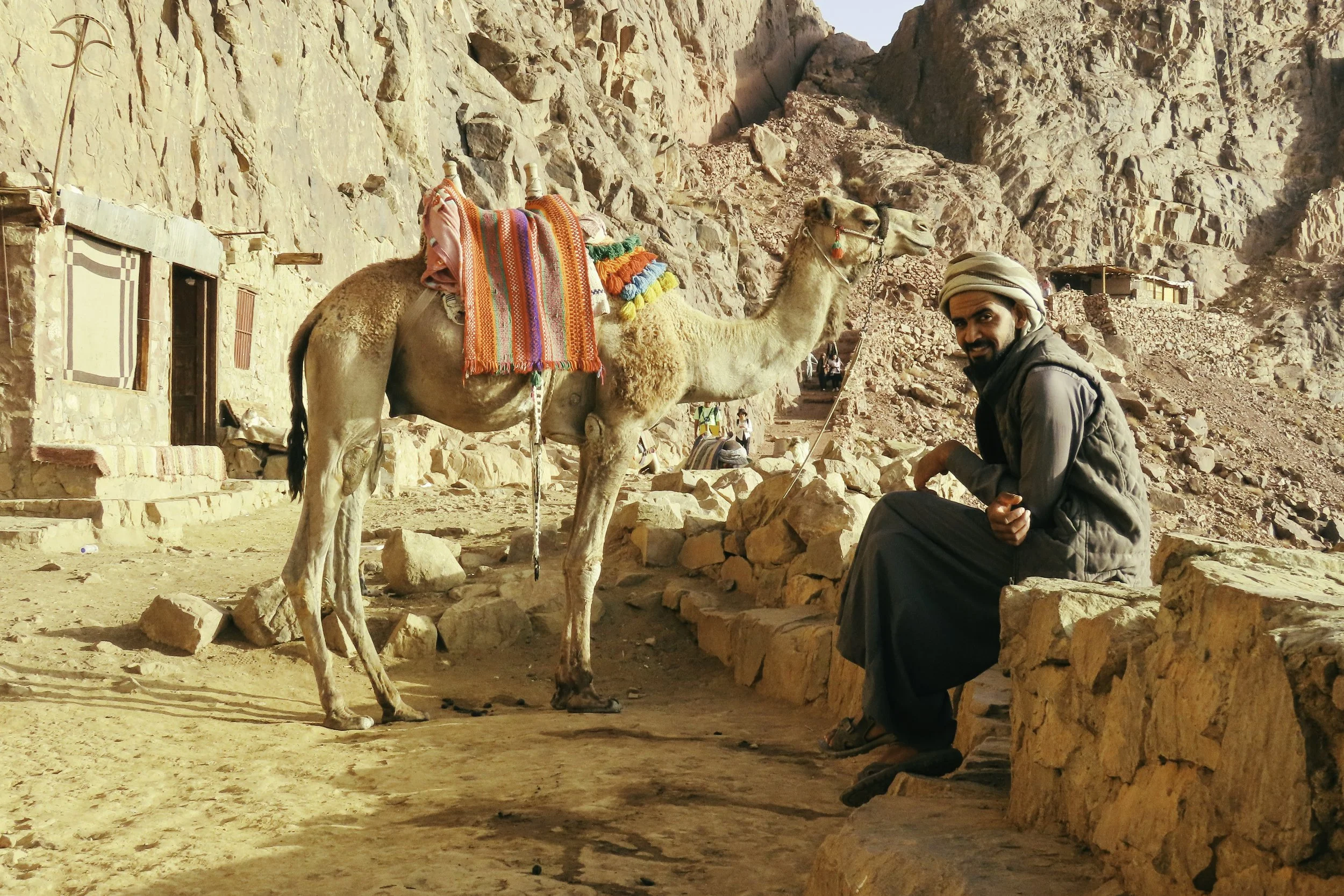 Uomo seduto su una panca di pietra accanto a un cammello nel deserto montuoso, con case di pietra sullo sfondo.