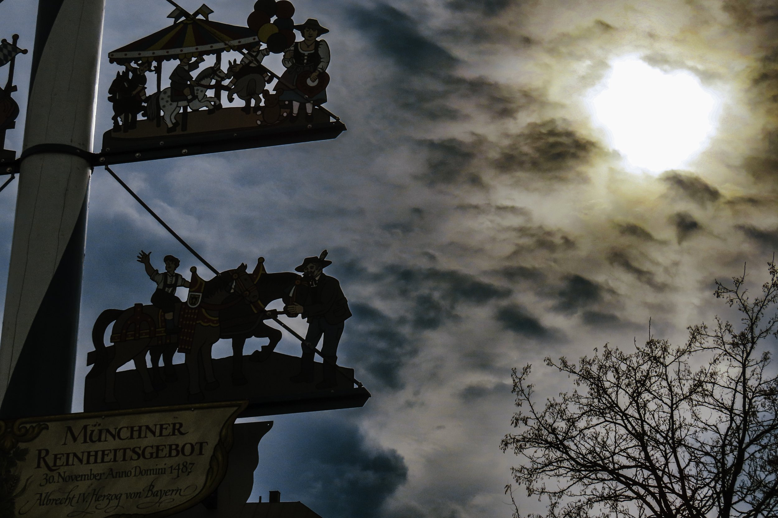 Mongi di legno decorativi con scene tradizionali sulla festa, sotto un cielo nuvoloso con il sole parzialmente coperto e alcuni rami di un albero nudo.
