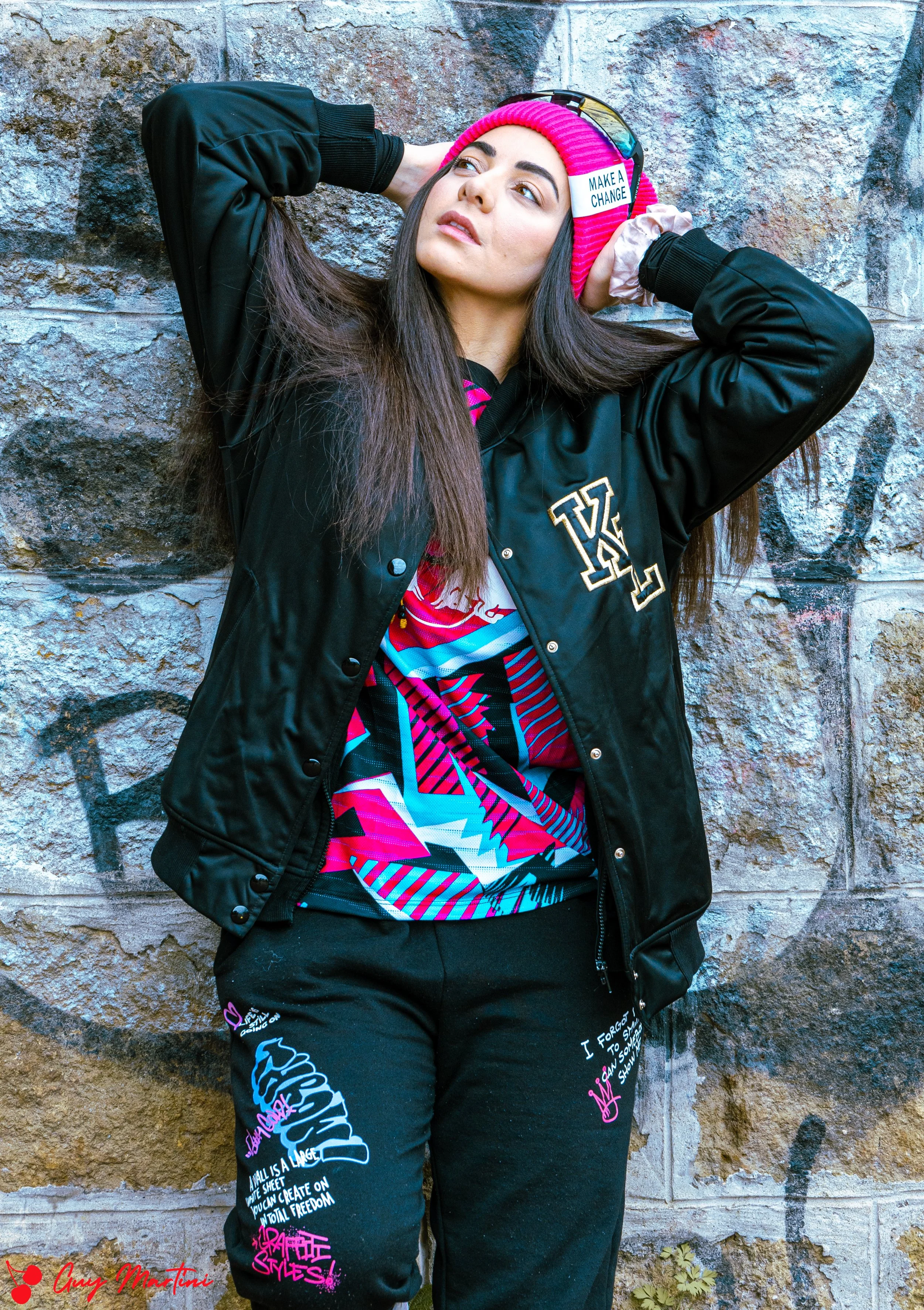Una ragazza indossa una giacca nera con artigli e pantaloni neri con scritte colorate, si trova davanti a un muro di pietra con graffiti, porta un berretto rosa con un adesivo che dice 'Make a Change'.
