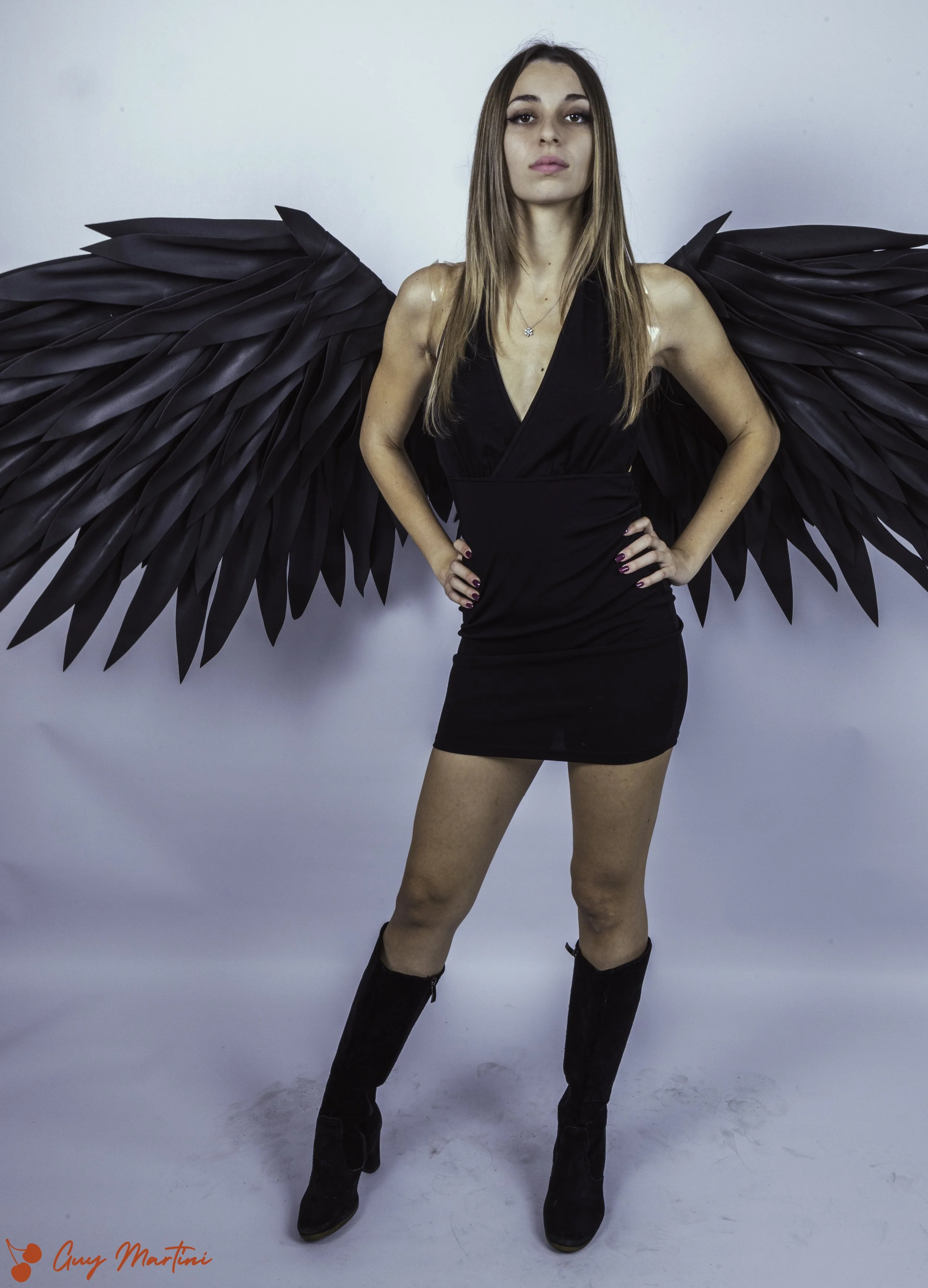 Donna giovane con ali nere di plastica, vestita con un abito nero cortissimo e stivali neri al ginocchio, posa con mano sui fianchi, sfondo bianco.