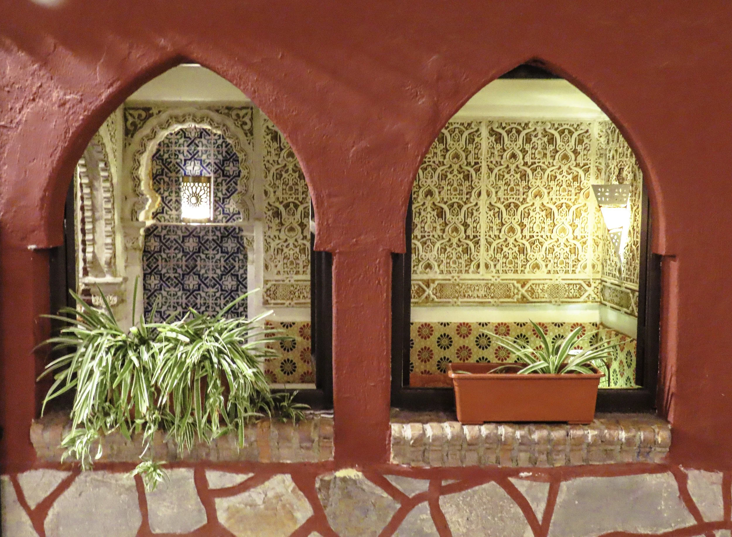 Vista di due finestrini con archi in stile arabo, decorati con piastrelle colorate e piante in vasi, in un ambiente con pareti decorate con motivi intricati.
