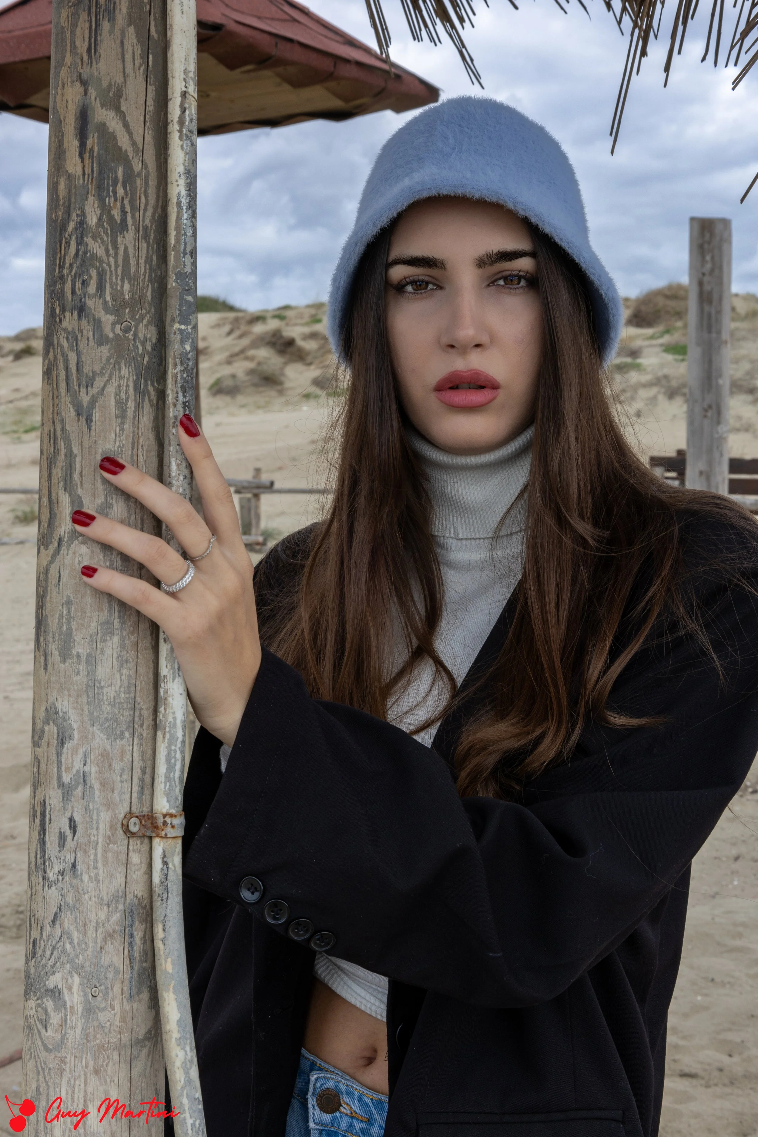 Ragazza con cappello azzurro, capelli castani, vestita con maglia bianca e giacca nera, in spiaggia con sabbia e strutture di legno in background.