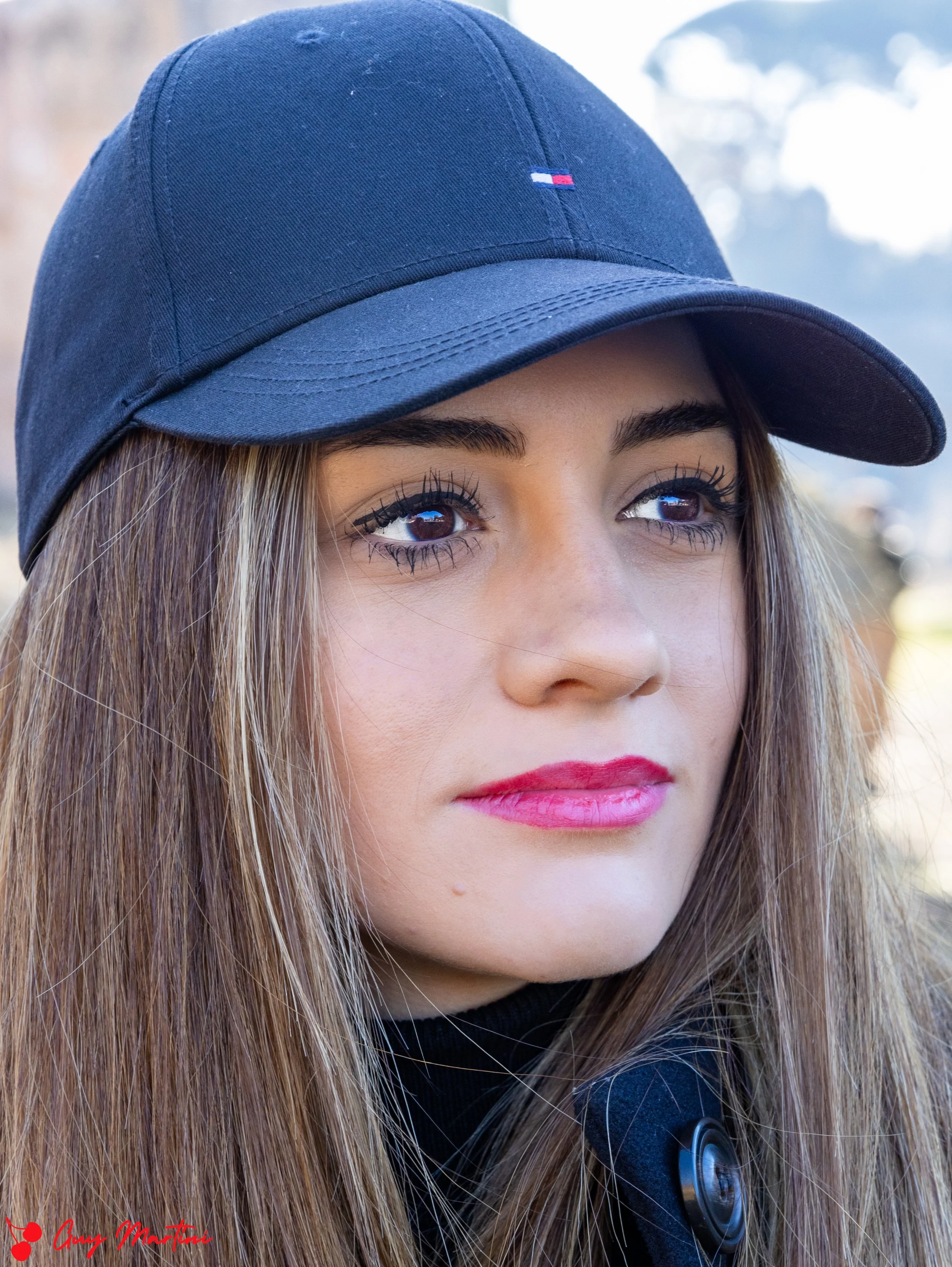 Una ragazza con cappello blu, capelli lunghi e chiari, trucco leggero e vestiti scuri, che guarda lontano
