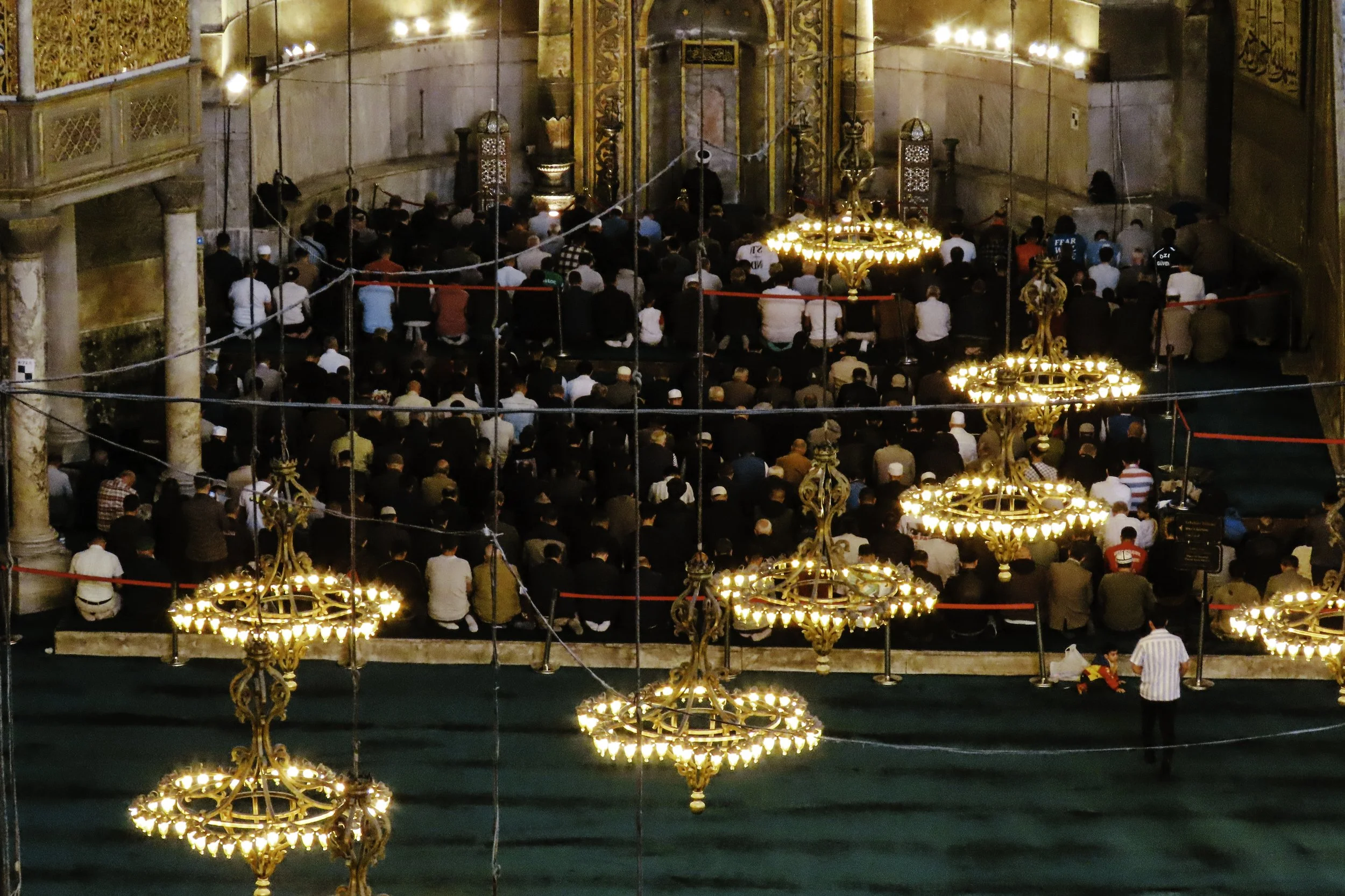 Fuori in una moschea, numerose persone pregano o si riuniscono per la preghiera, illuminate da grandi lampadari dorati e ghirlande di luci sospese nel soffitto.