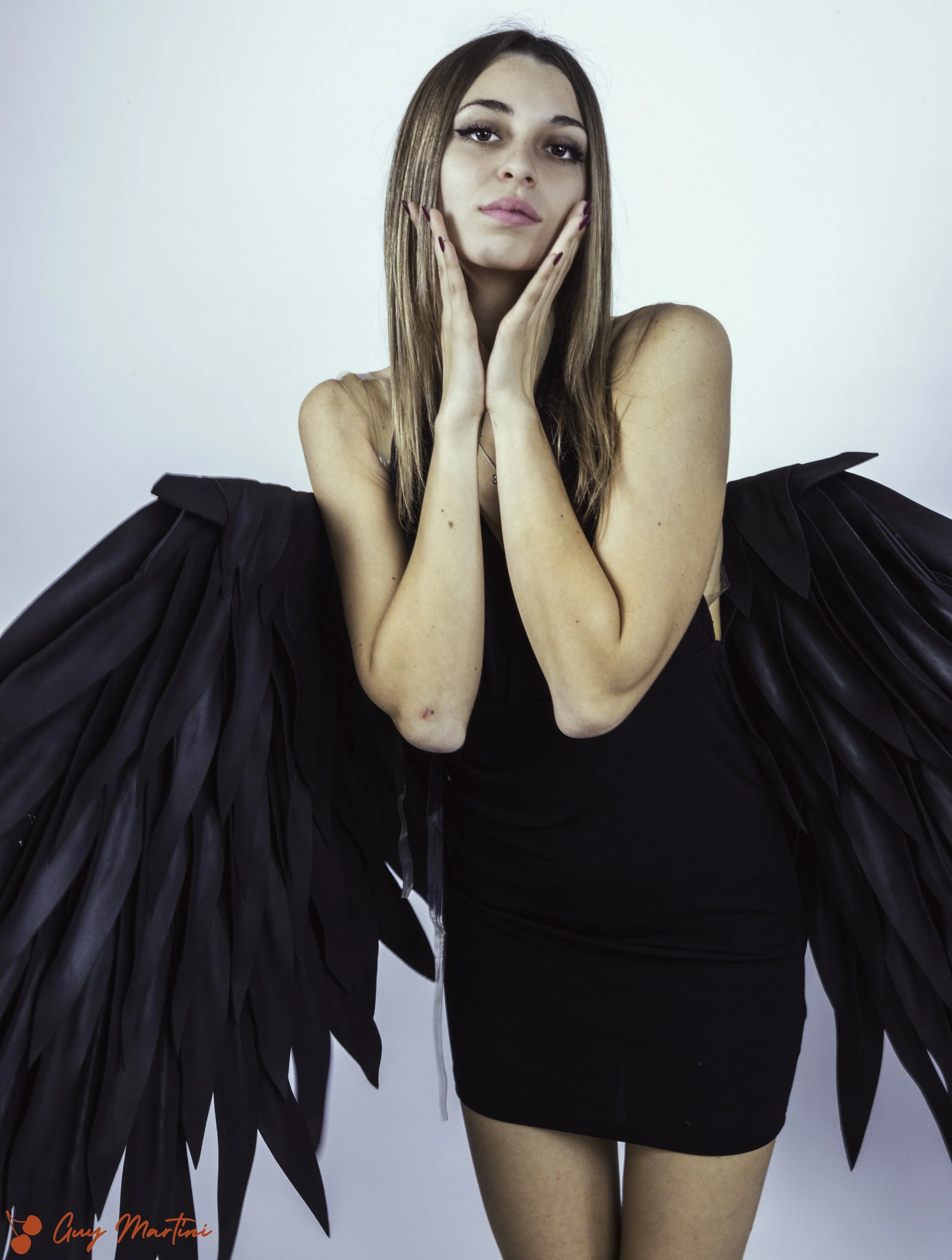 Una giovane donna con abiti neri, con alamari di piume nere sulle spalle, si trova davanti a uno sfondo bianco. La donna ha i capelli castani lunghi e lisci, e sta tenendo le mani sul viso con un’espressione serena e sicura.