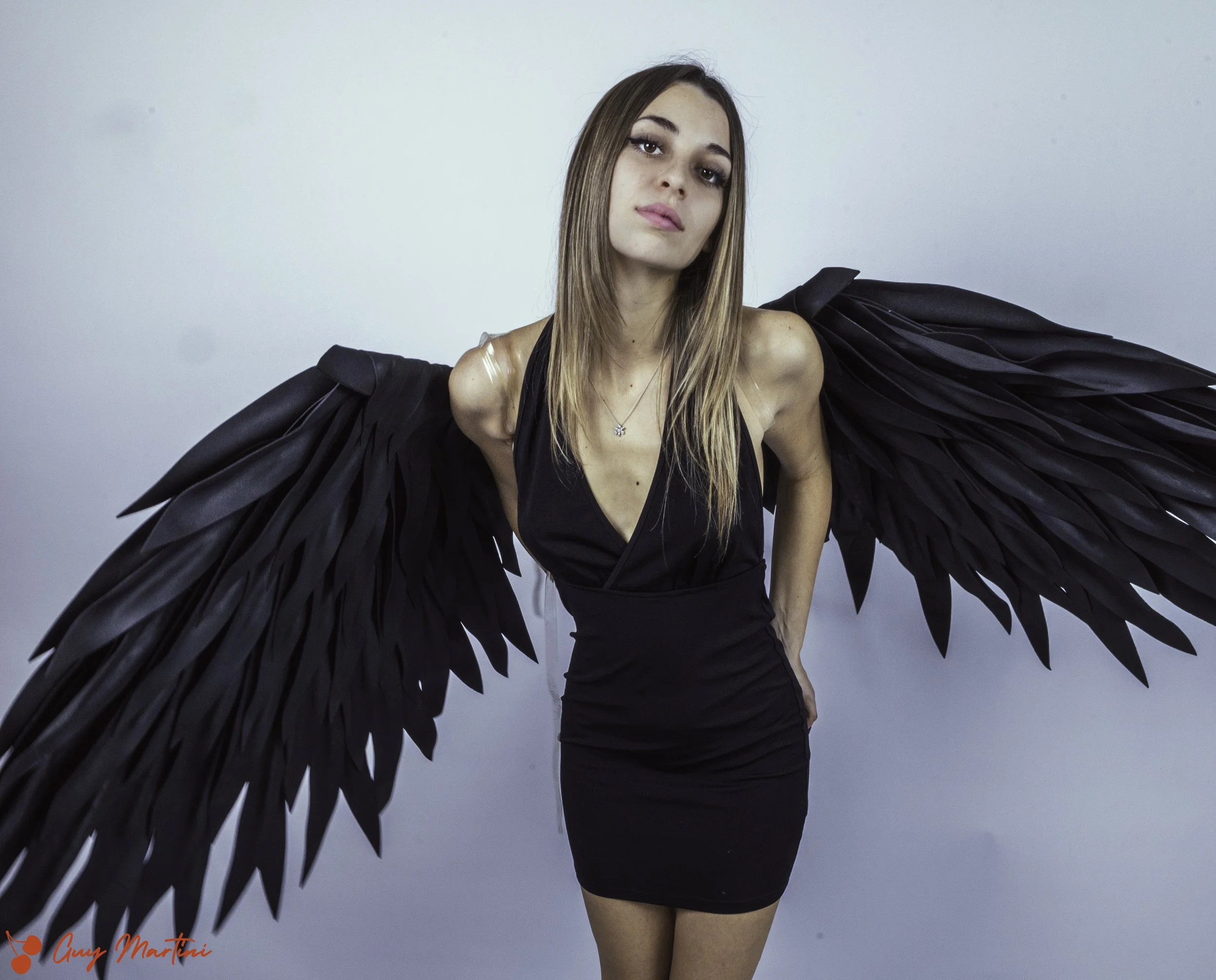 Una donna con ali nere di piume indossa un vestito nero e si trova davanti a uno sfondo grigio