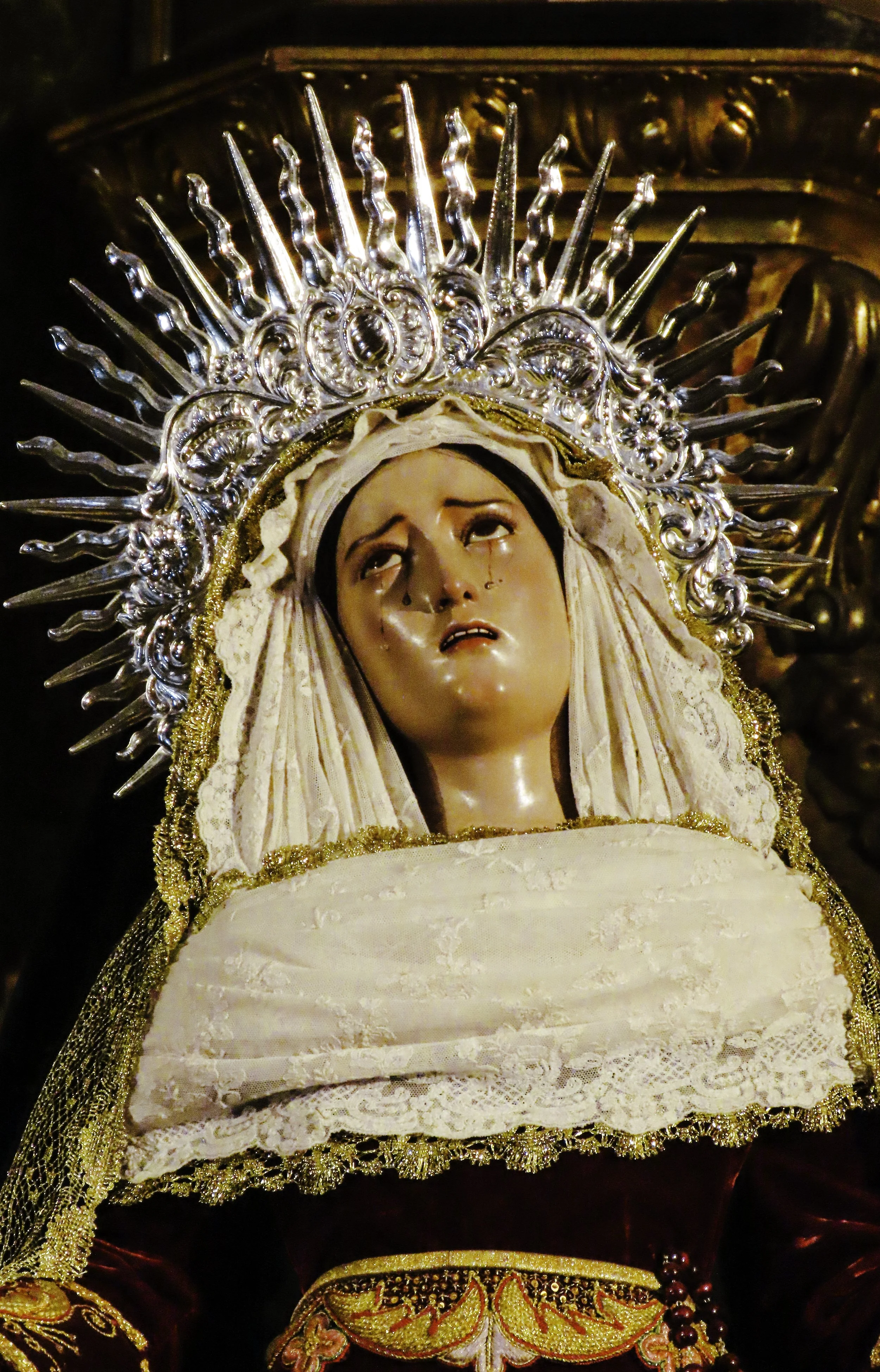 Statua religiosa di Maria con espressione triste, indossa un mantello ricamato e una corona decorata con raggi, in un ambiente sacro.