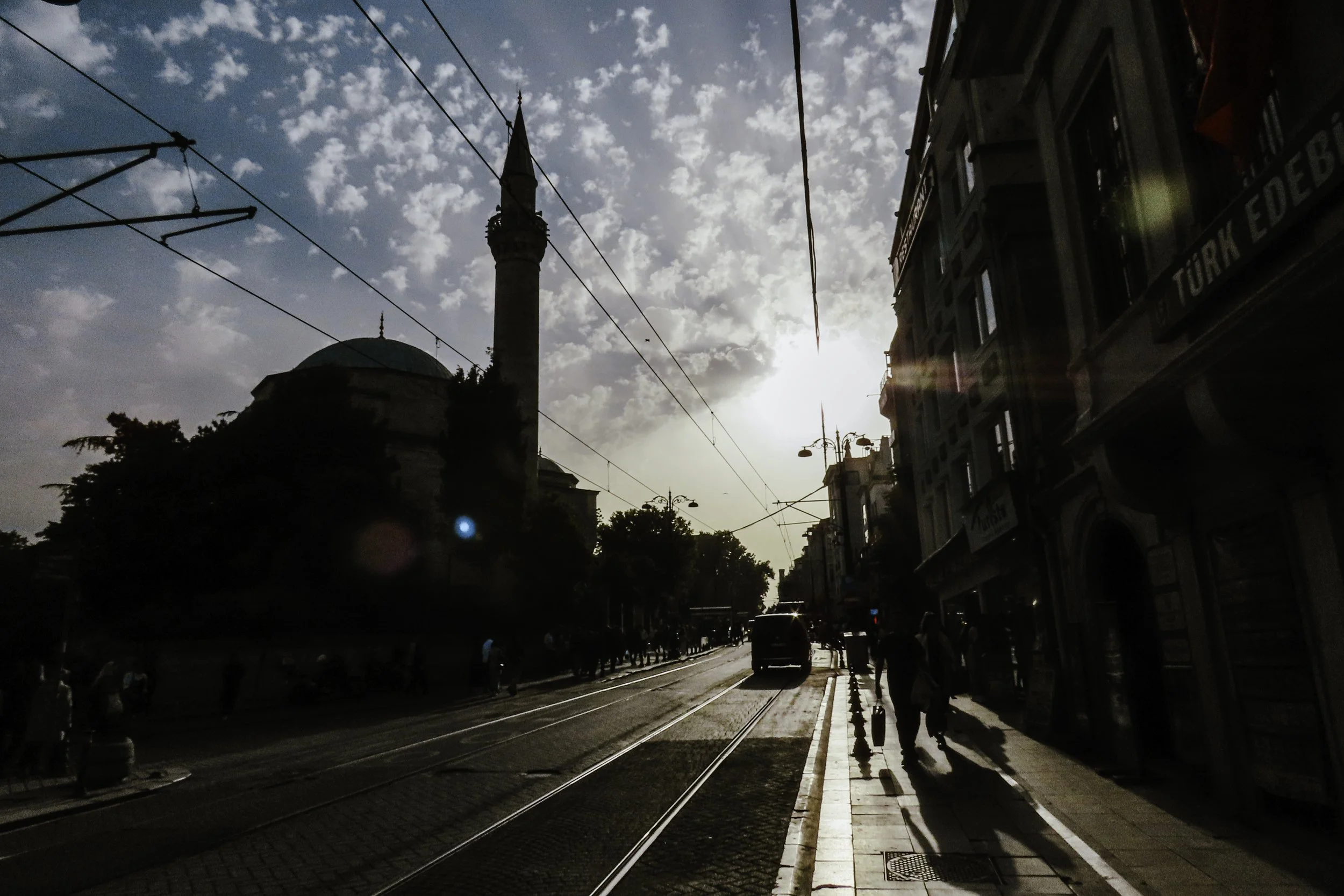 Una strada cittadina con tram e pedoni, con un minareto e una moschea sullo sfondo, illuminata dal sole al tramonto