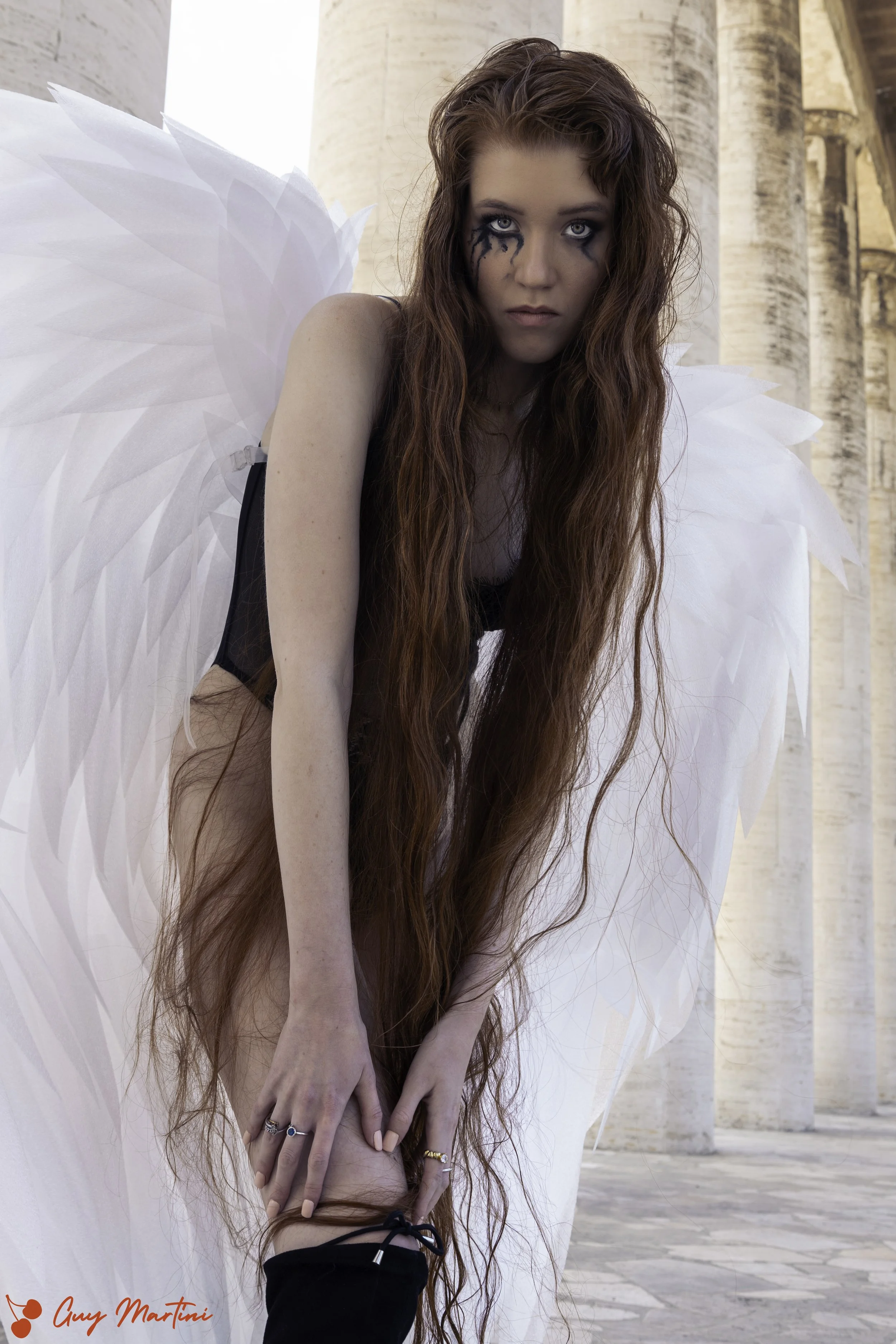 Una donna con lunghi capelli ricci, trucco pesante sugli occhi, indossa un costume da angelo con grandi ali bianche, si trova di fronte a una struttura in pietra.