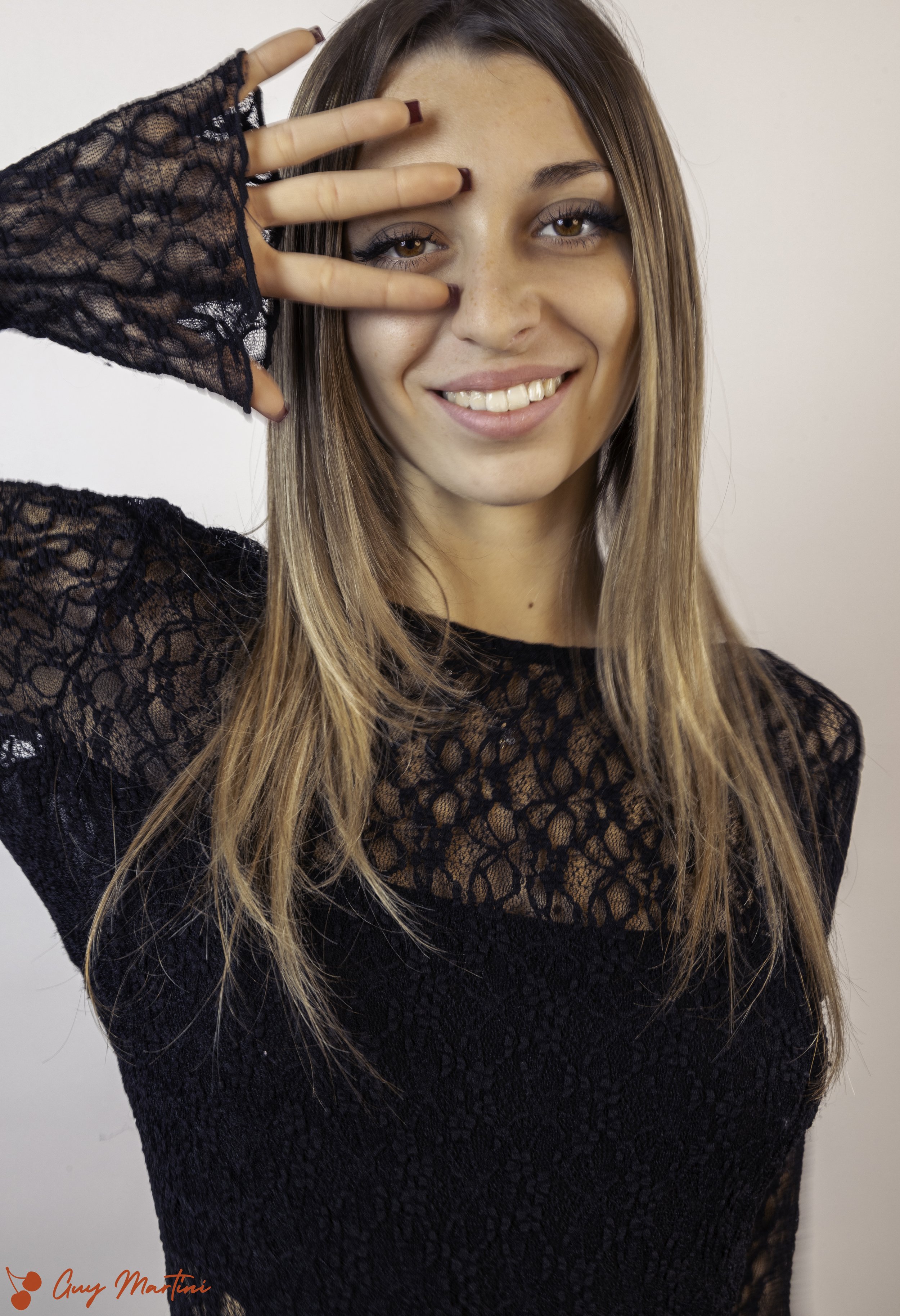 Ragazza sorridente che indossa un maglione nero in pizzo, fa un gesto con tre dita vicino alla fronte e guarda verso la fotocamera.