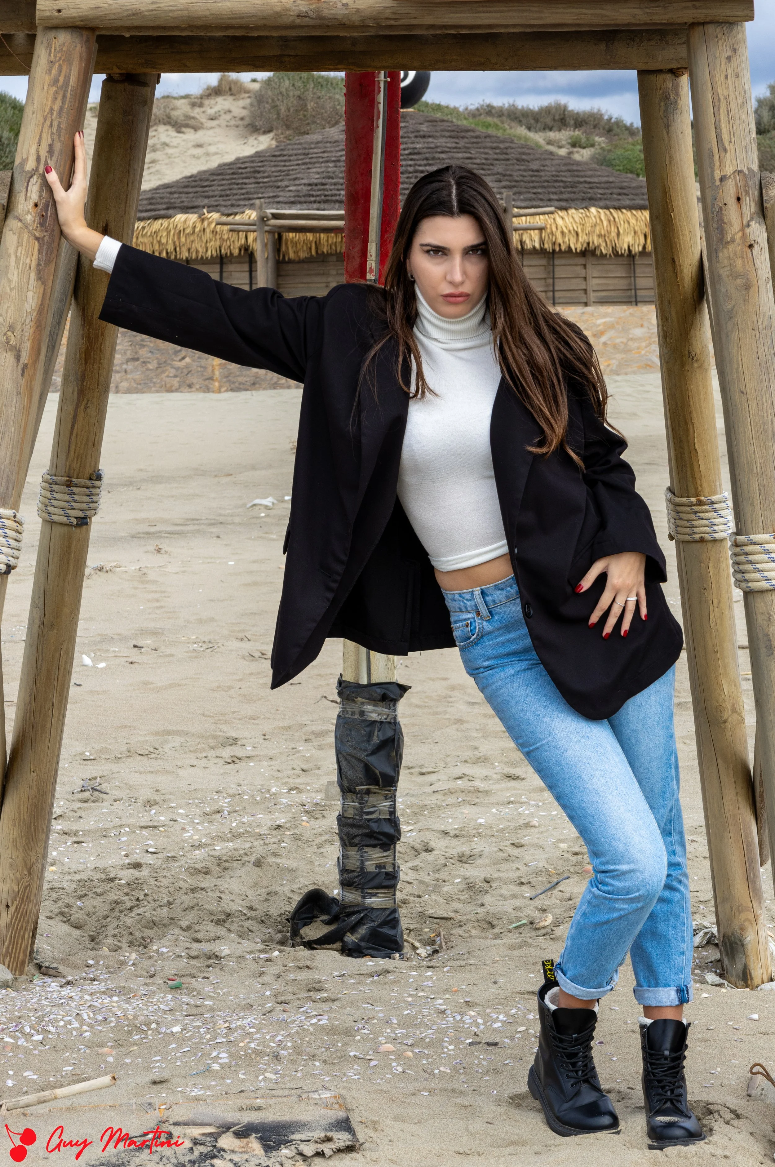 Donna che posa sotto una struttura di legno sulla spiaggia, indossa una giacca nera, felpa bianca, jeans e stivali neri, con sfondo di case di paglia e sabbia.