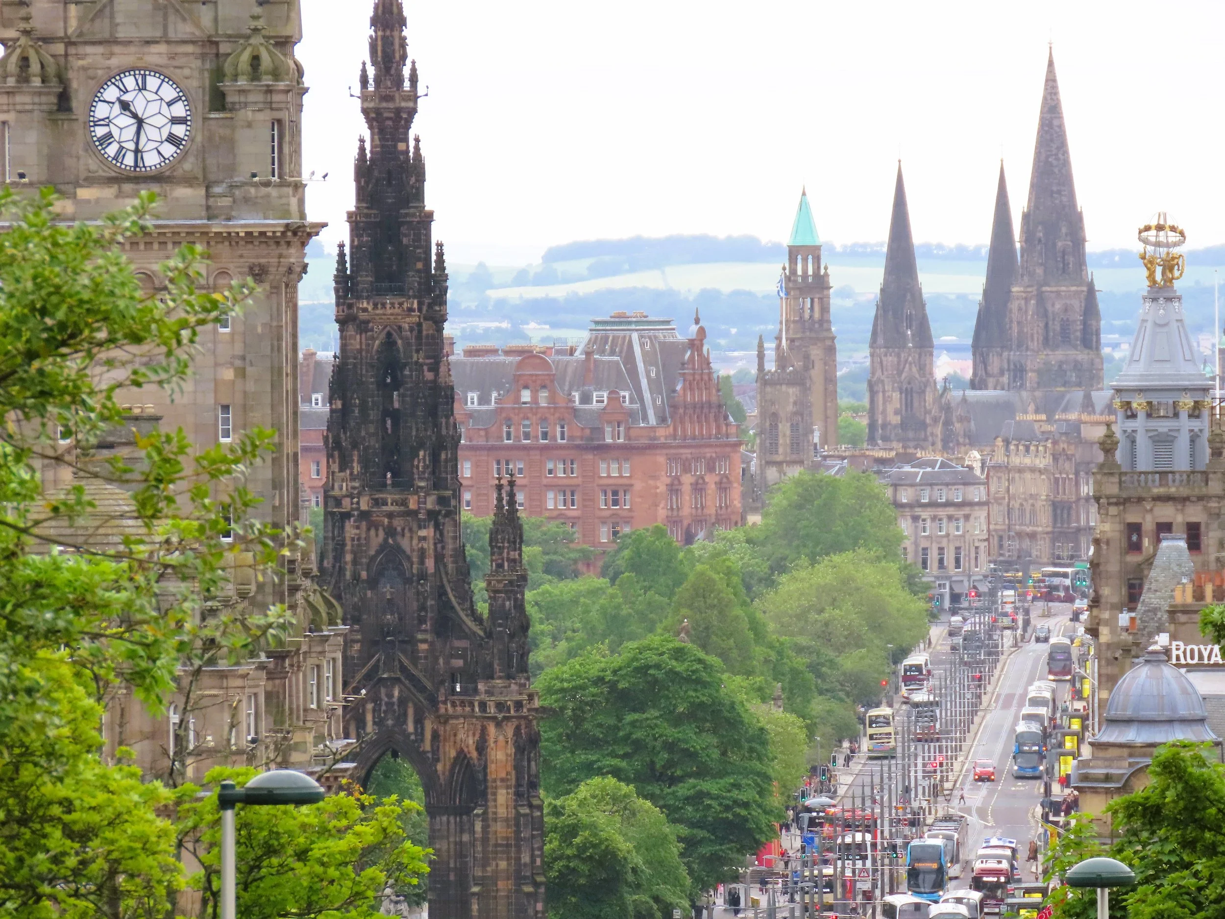 Veduta di una città con edifici storici e chiese con torri alte, affollata di automobili e autobus, con alberi verdi lungo la strada e un cielo nuvoloso.
