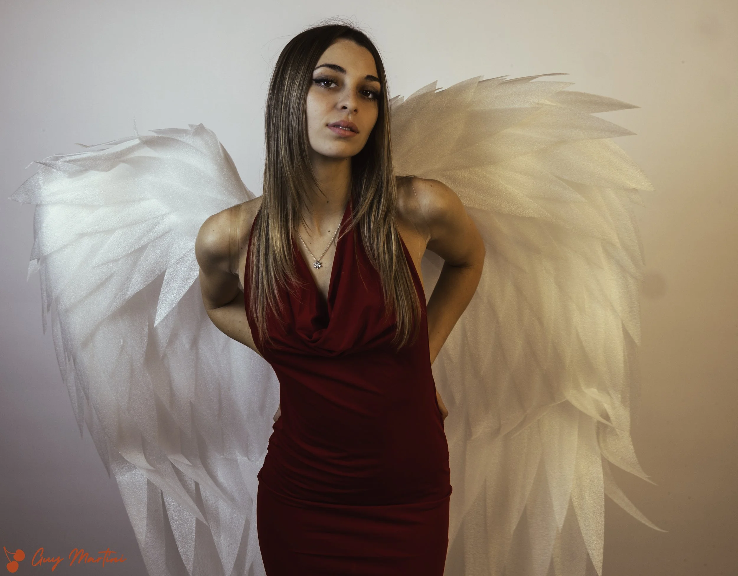 Una donna con lunghi capelli castani, vestita con un abito rosso scuro, che indossa ali di angelo bianche e piumate. È posizionata di fronte a uno sfondo neutro e guarda leggermente a sinistra. Nell'angolo in basso a sinistra si legge una firma rossa