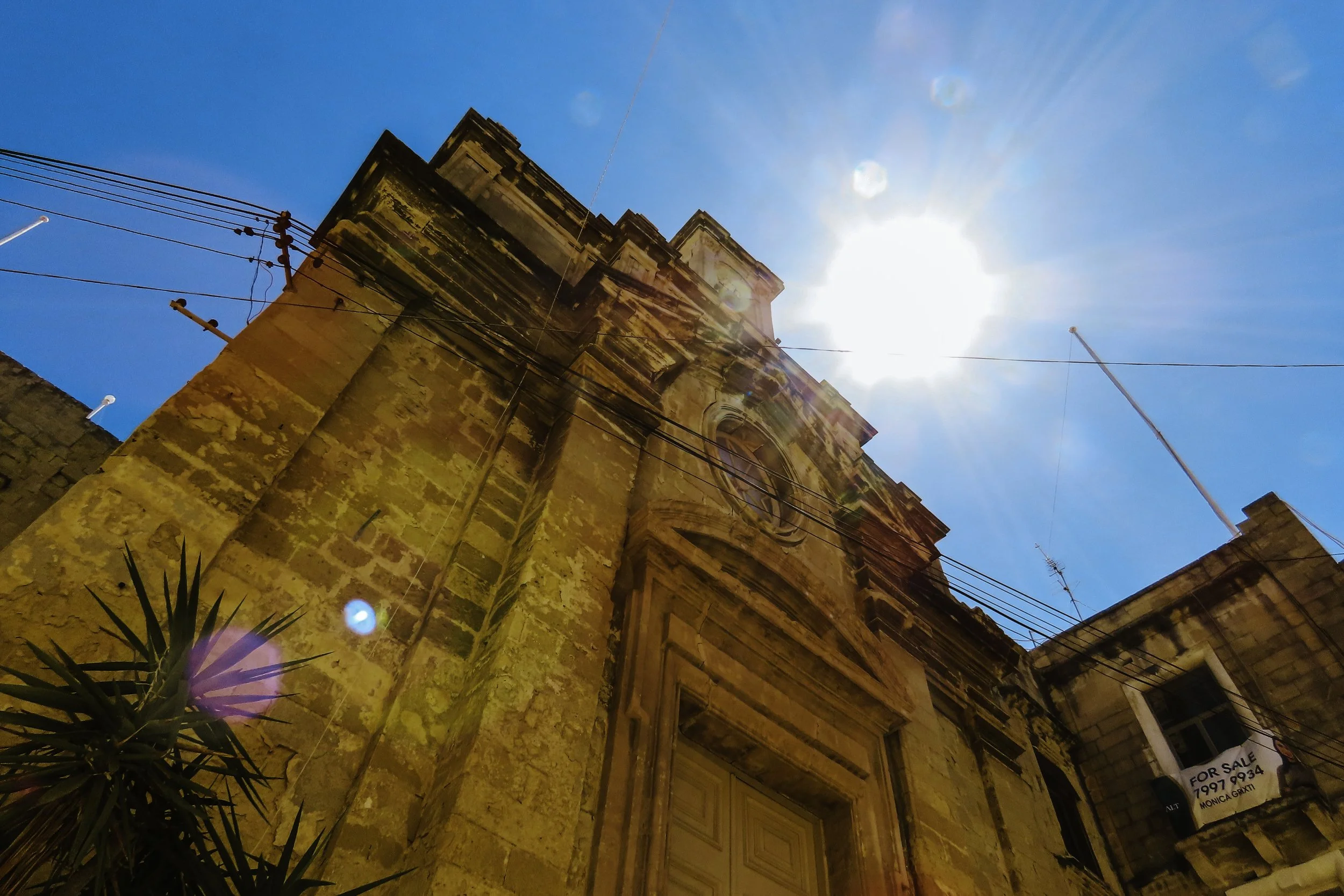 Vista di un edificio storico in pietra, con un grande portale e un orologio, sotto un cielo blu con il sole splendente e raggi che irradiato.