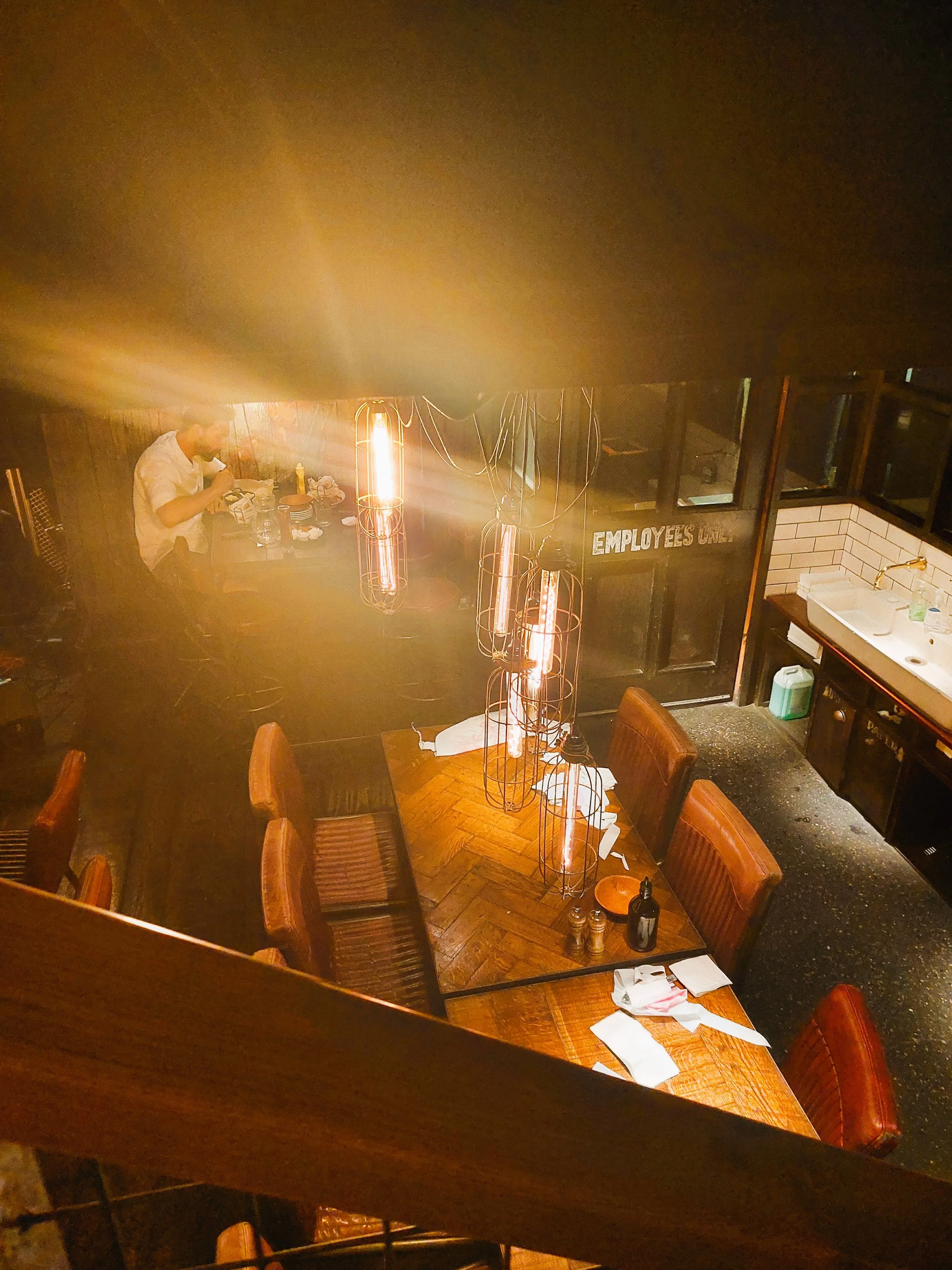 Interno di un ristorante con un tavolo in legno, sedie in pelle marrone, luci a lampada industriale, pareti di mattoni bianchi e un lavandino sullo sfondo.