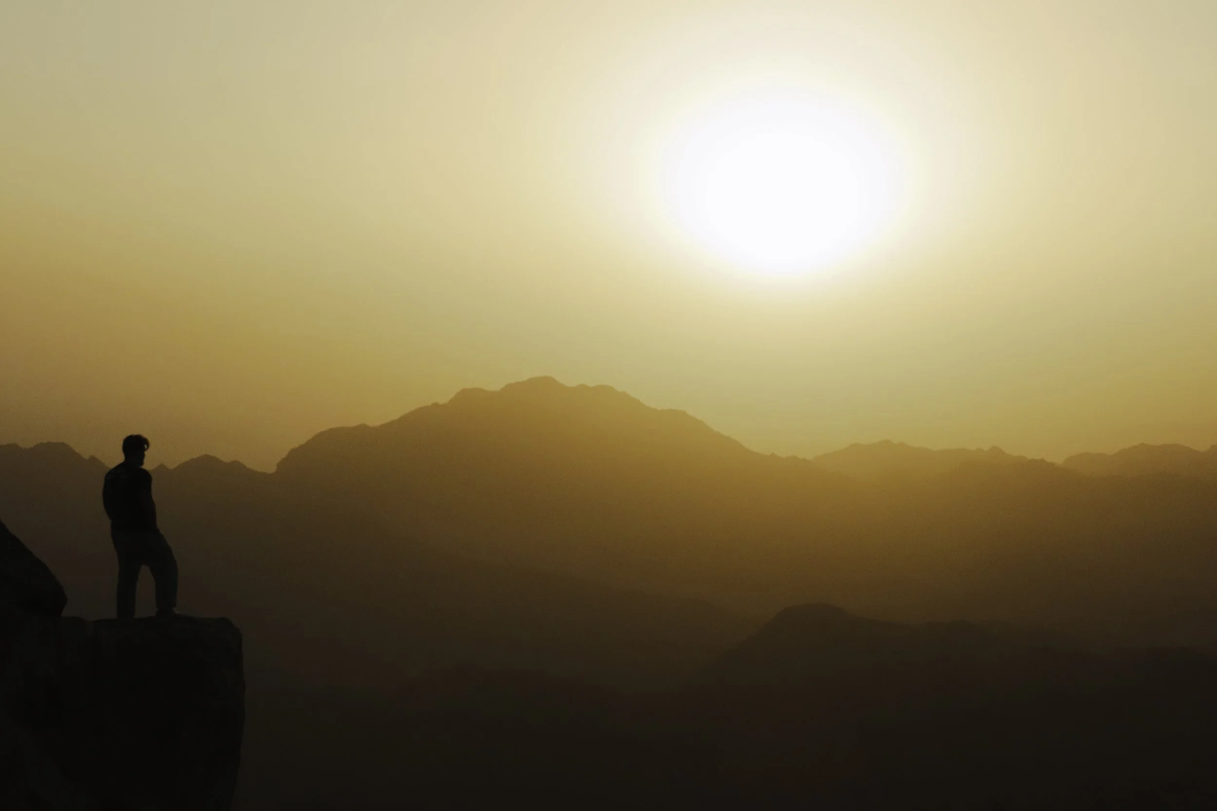 Persona in piedi su una roccia in un paesaggio montano al tramonto o all'alba, con il sole che splende sullo sfondo e le montagne all'orizzonte.