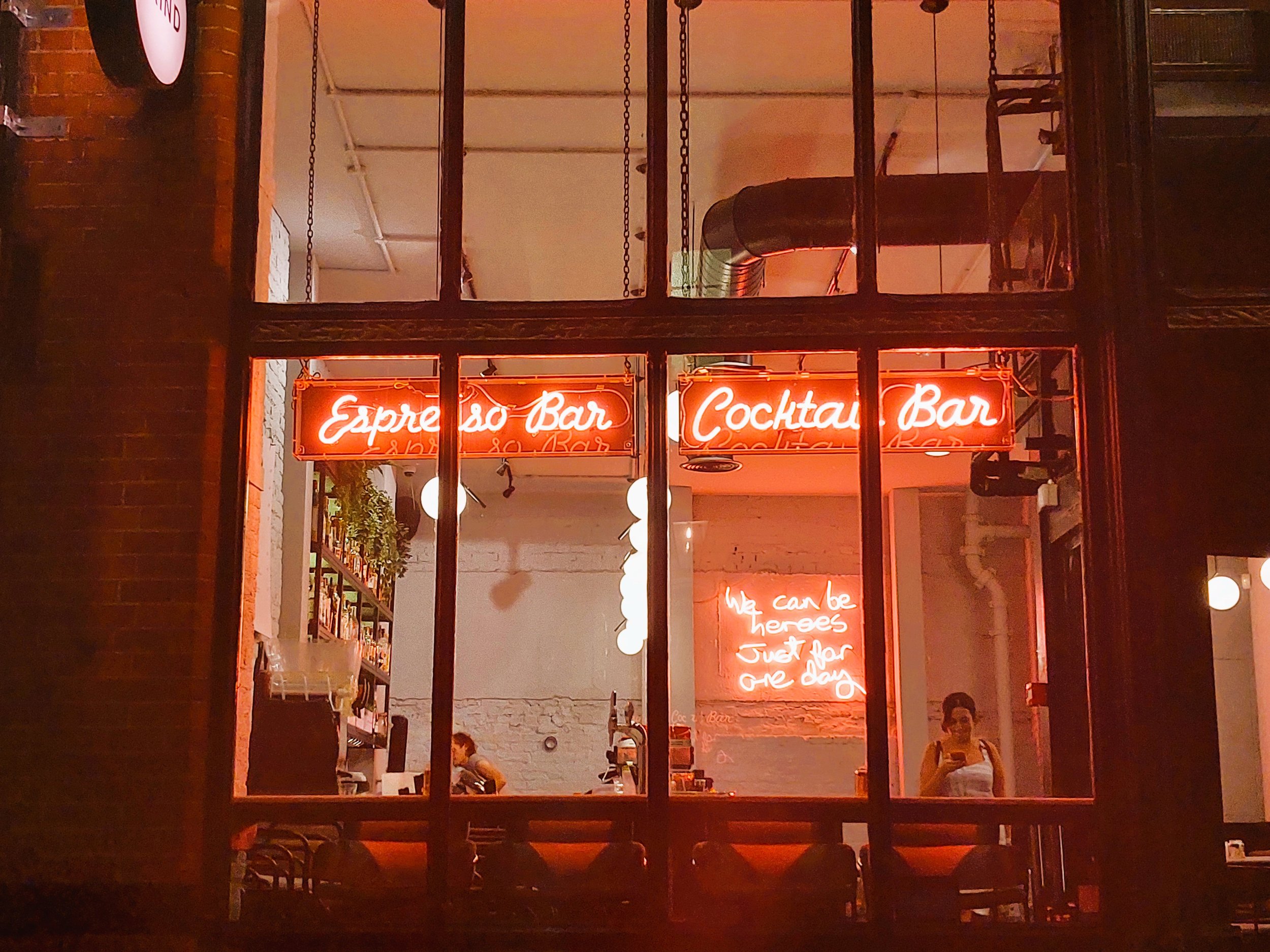 Interno di un bar con luci al neon rosse che dice 'Esprresso Bar' e 'Cocktail Bar'. Una donna prende un selfie e due persone sono al bancone.