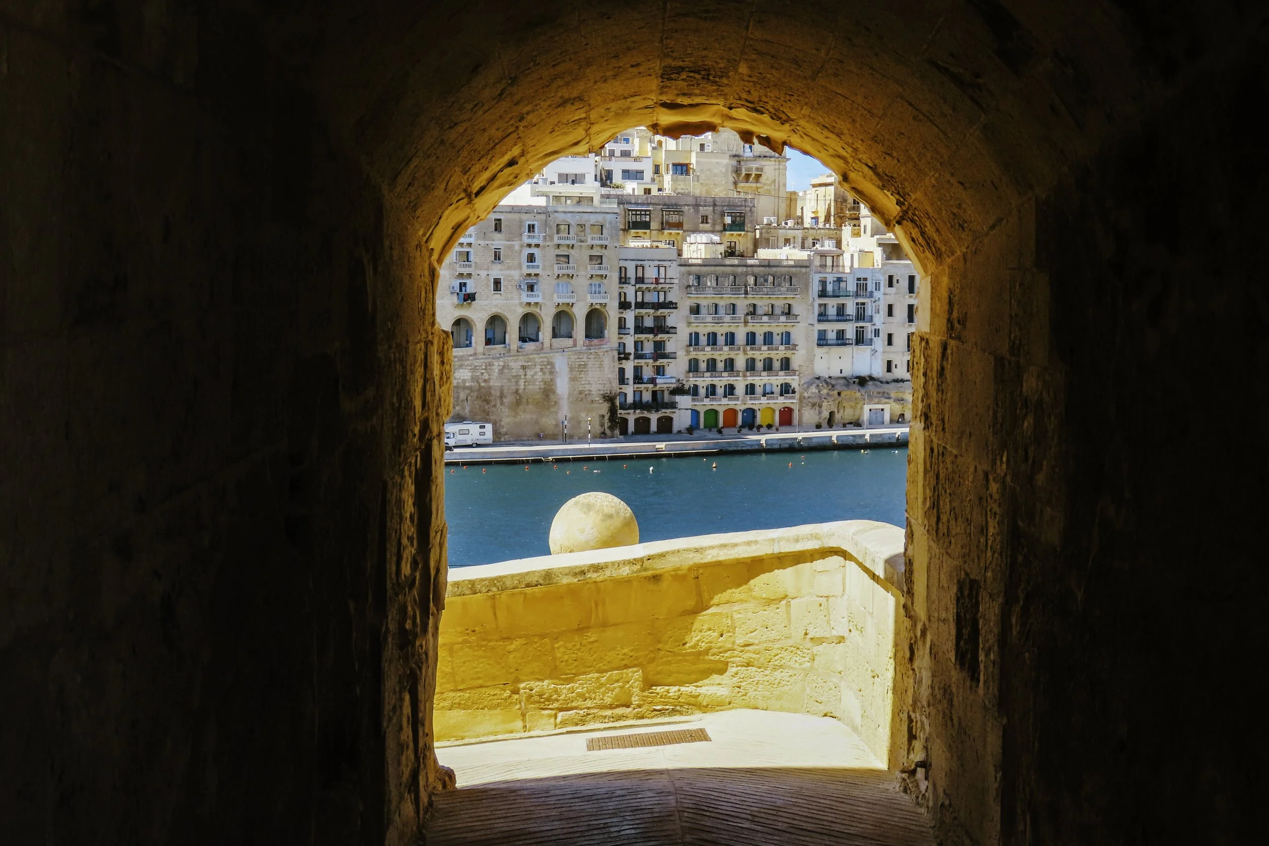Vista panoramica di un porto con edifici colorati e una torre di pietra, vista attraverso un'apertura di una fortezza o mura antiche, con il mare sullo sfondo.