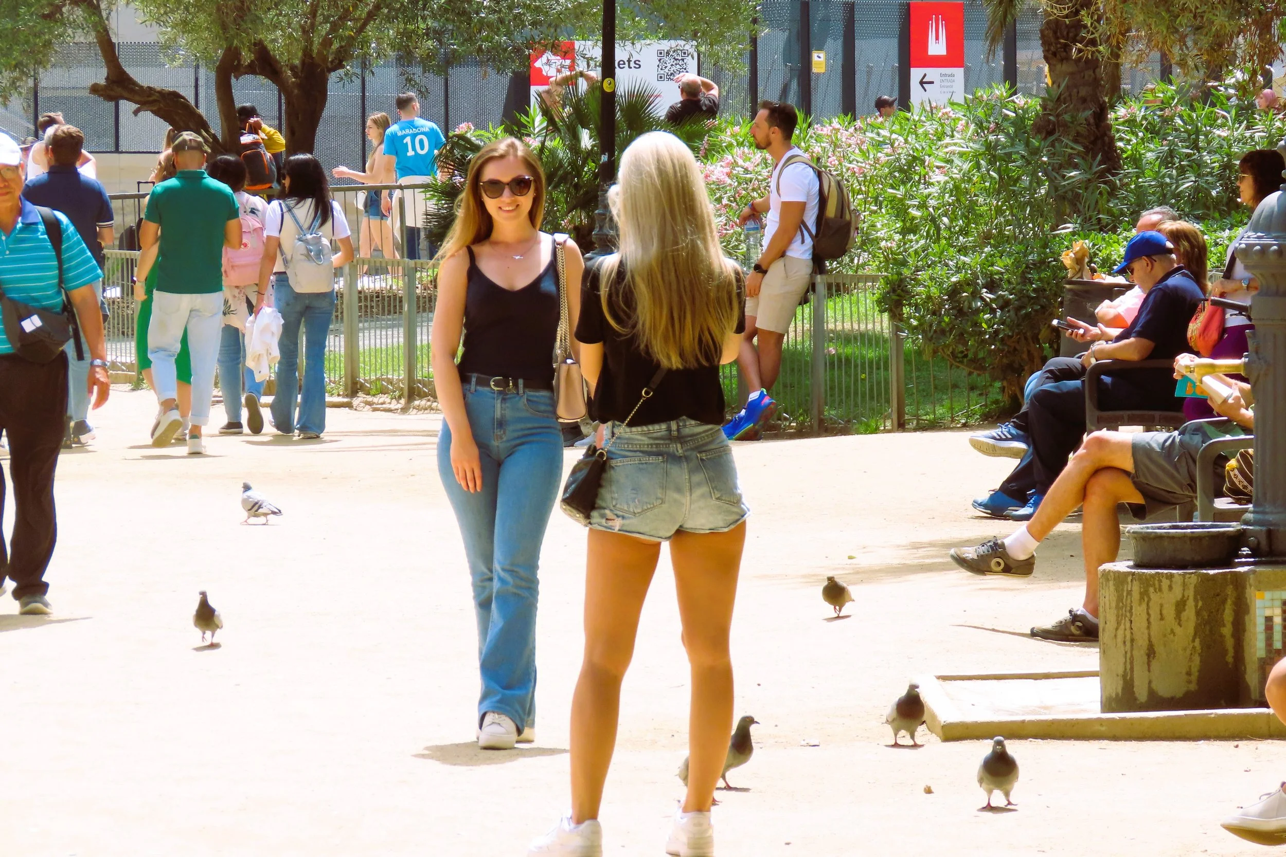 Persone che passeggiano e si rilassano in un parco, con due ragazze in primo piano che parlano tra loro, all'aperto in un giorno di sole.