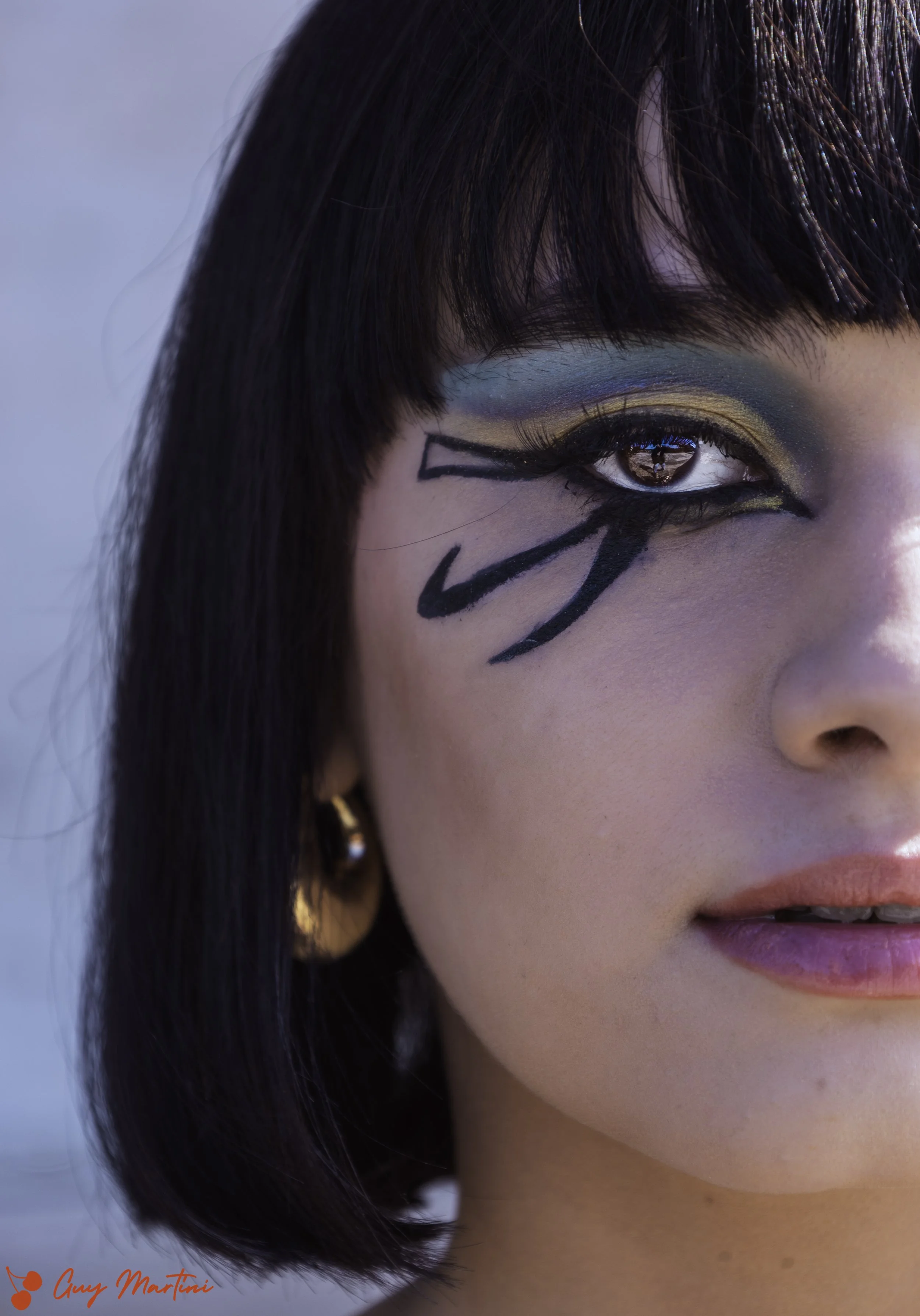 Primo piano di una donna con trucco artistico sugli occhi, con eyeliner nero e ombreggiatura blu e oro, con dettagli di calligrafia nera sul lato del viso vicino all'occhio.