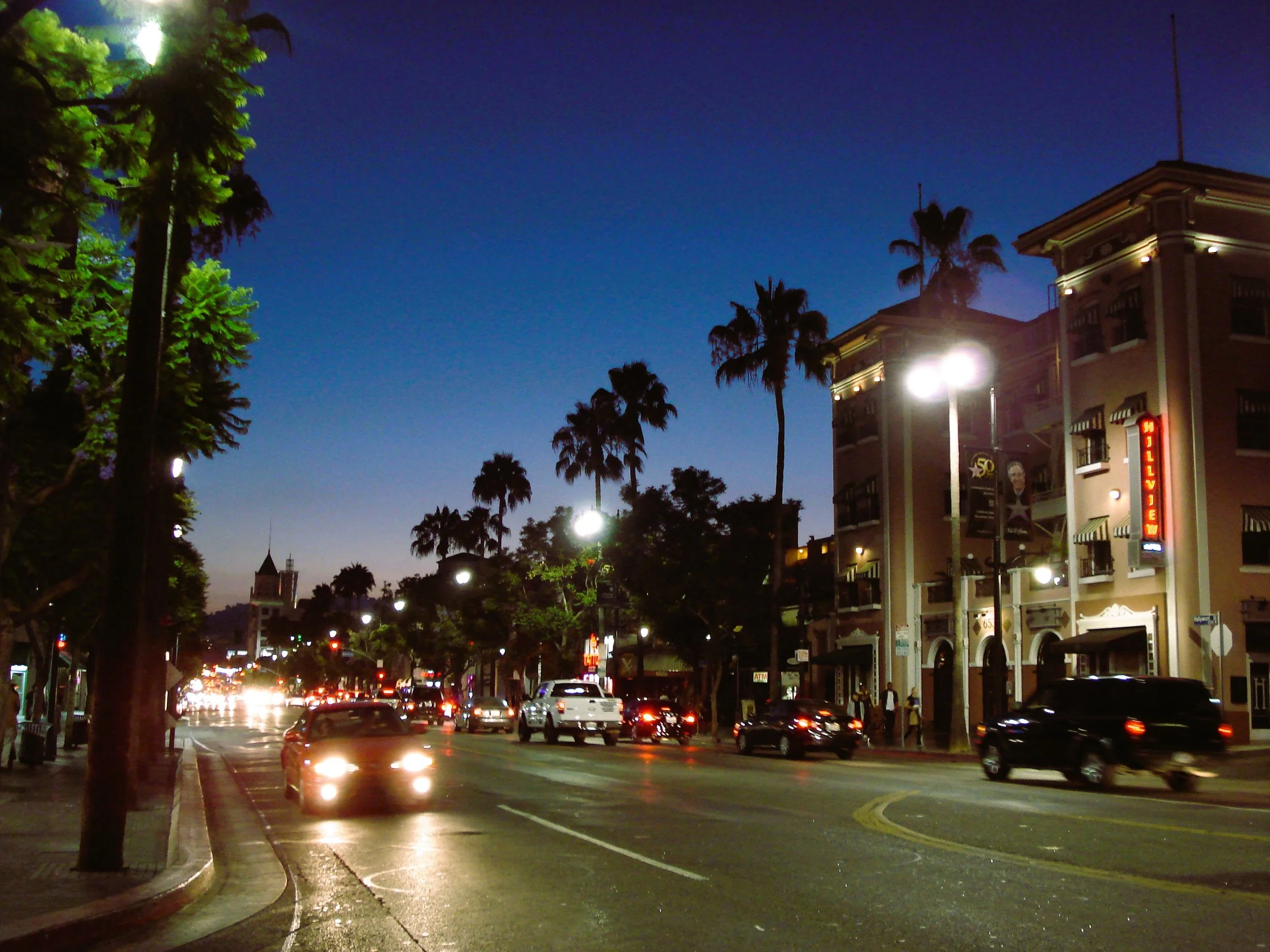 Una strada cittadina con negozi e alberi al tramonto, luci accese e auto in movimento.