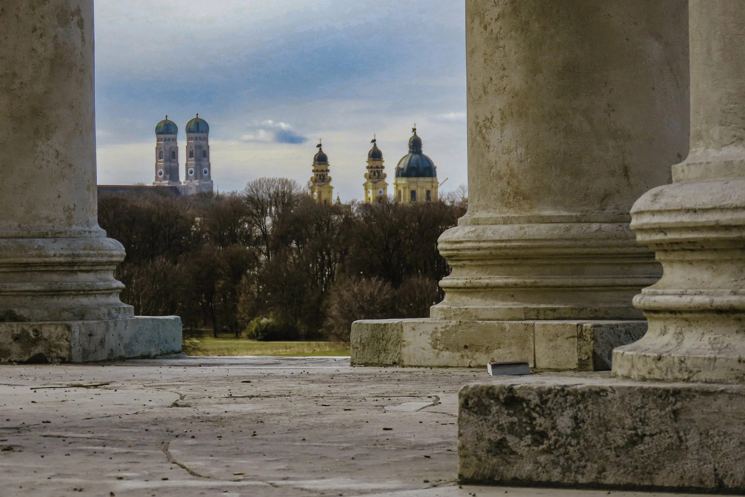 Vista attraverso colonne di pietra che mostra un parco con alberi e chiese con cupole blu e torri con cupole verdi sullo sfondo, cielo leggermente nuvoloso.