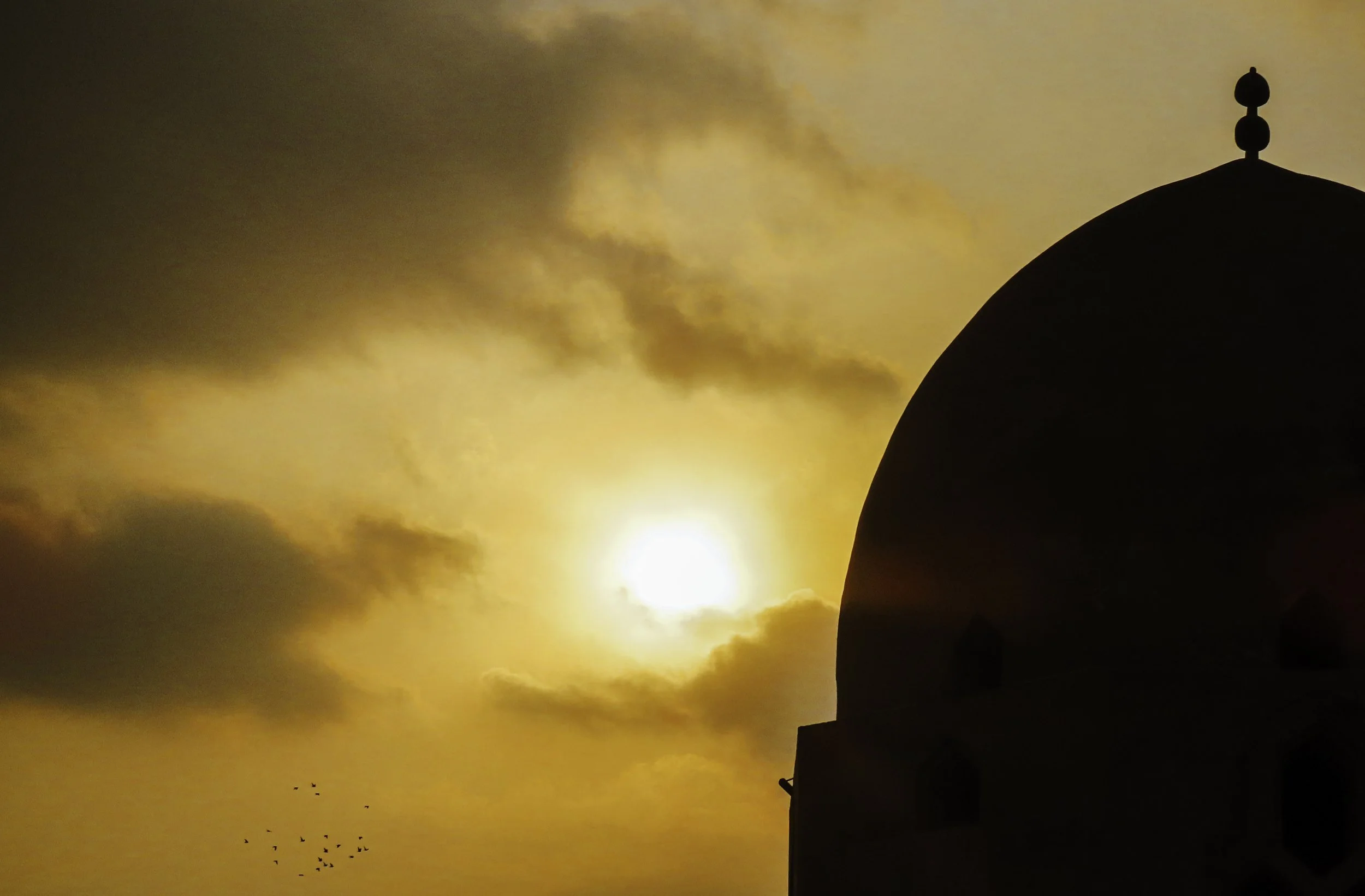 Siluetta di una cupola di un edificio religioso contro un cielo al tramonto con il sole tra le nuvole e un gruppo di uccelli volando in basso.