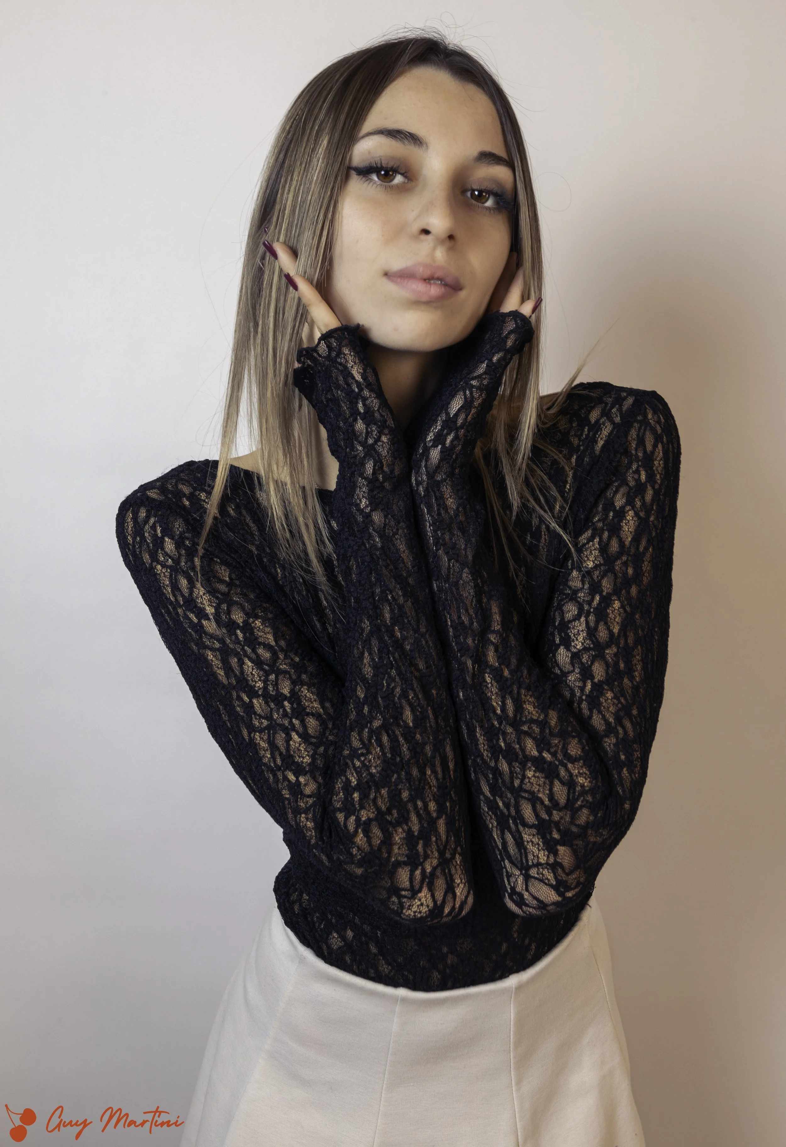 Ragazza con lunghi capelli castani, indossa una maglia nera in pizzo trasparente, posa con le mani sul volto, sfondo chiaro.