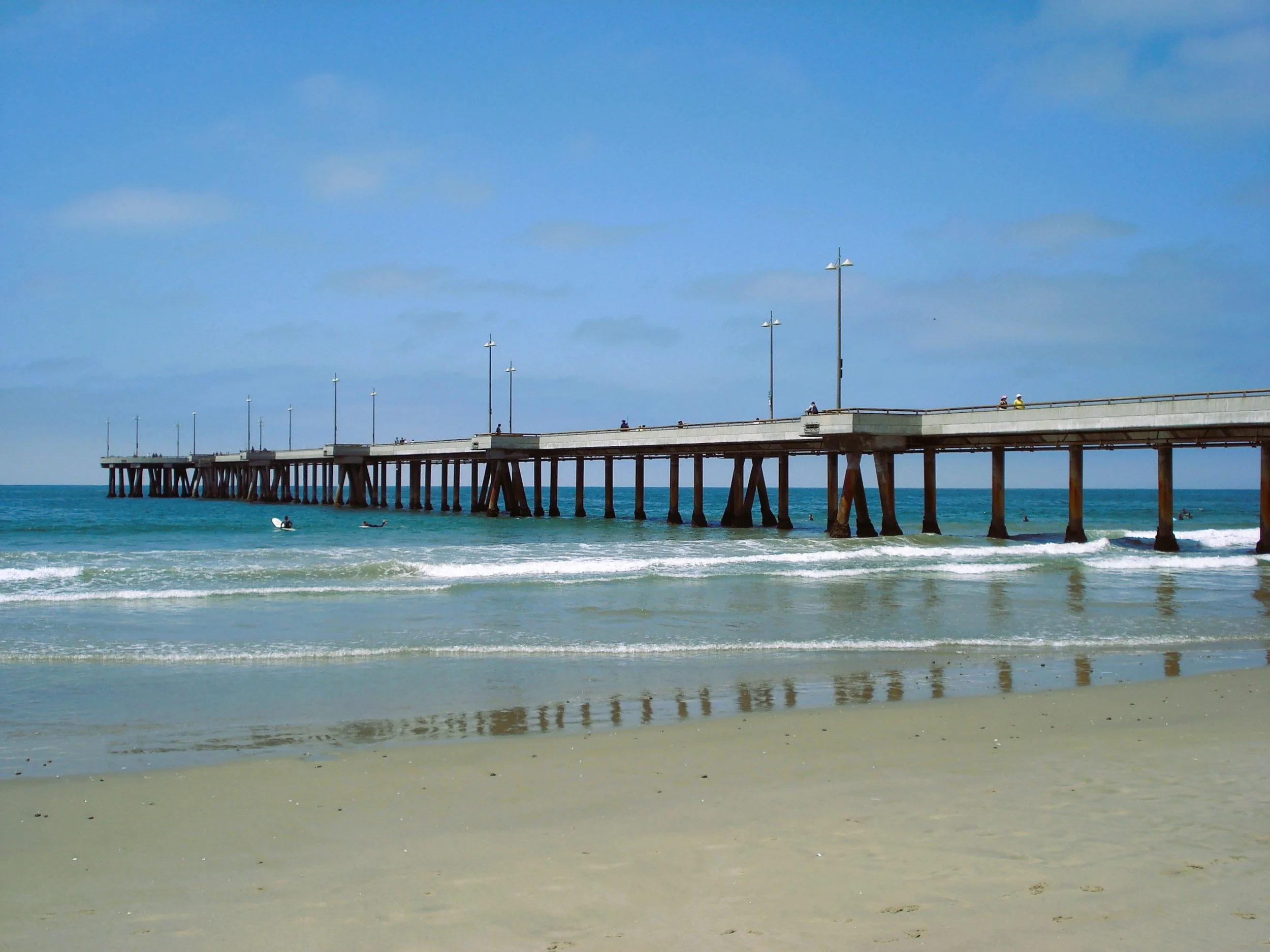 Spiaggia con mare calmo, pontile di legno e alcune persone che si divertono in acqua.