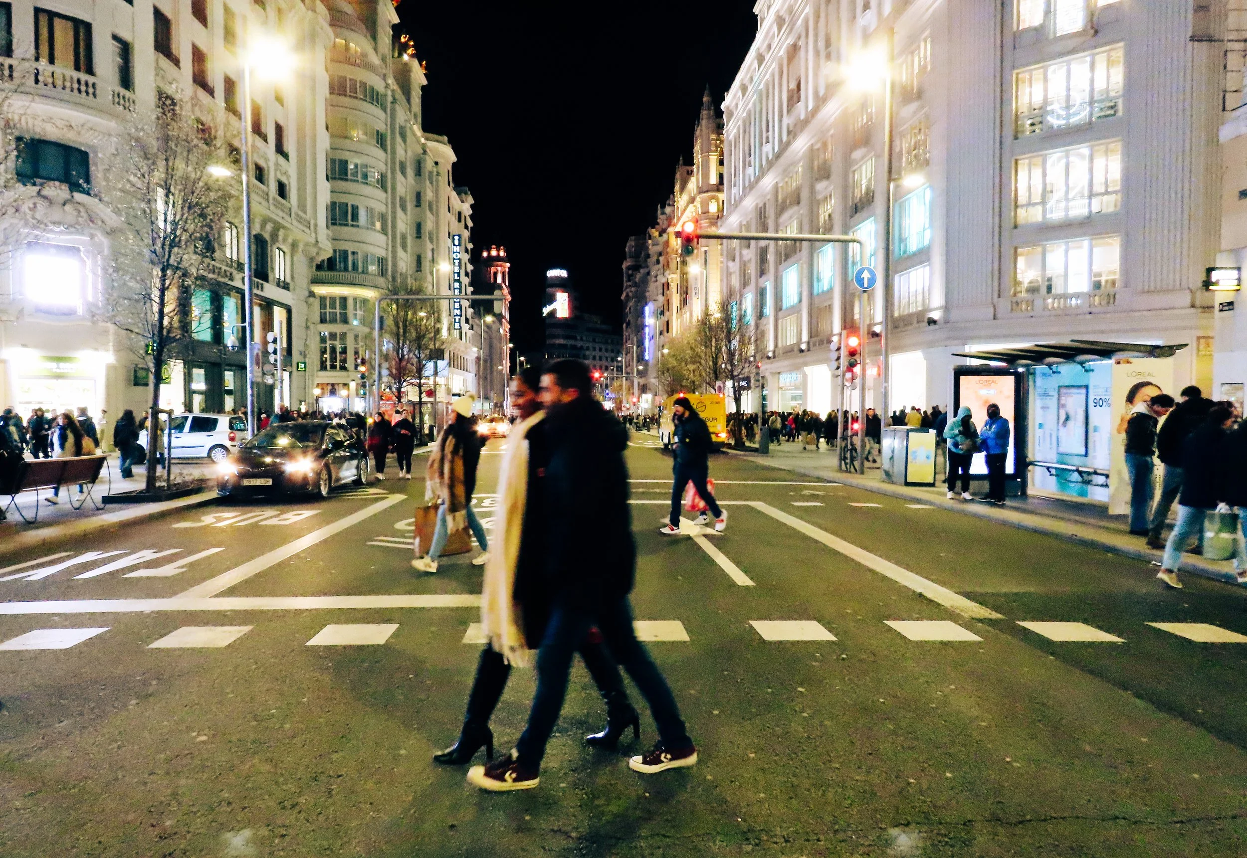 Una strada affollata in città di notte, con persone che attraversano le strisce pedonali e edifici illuminati ai lati.