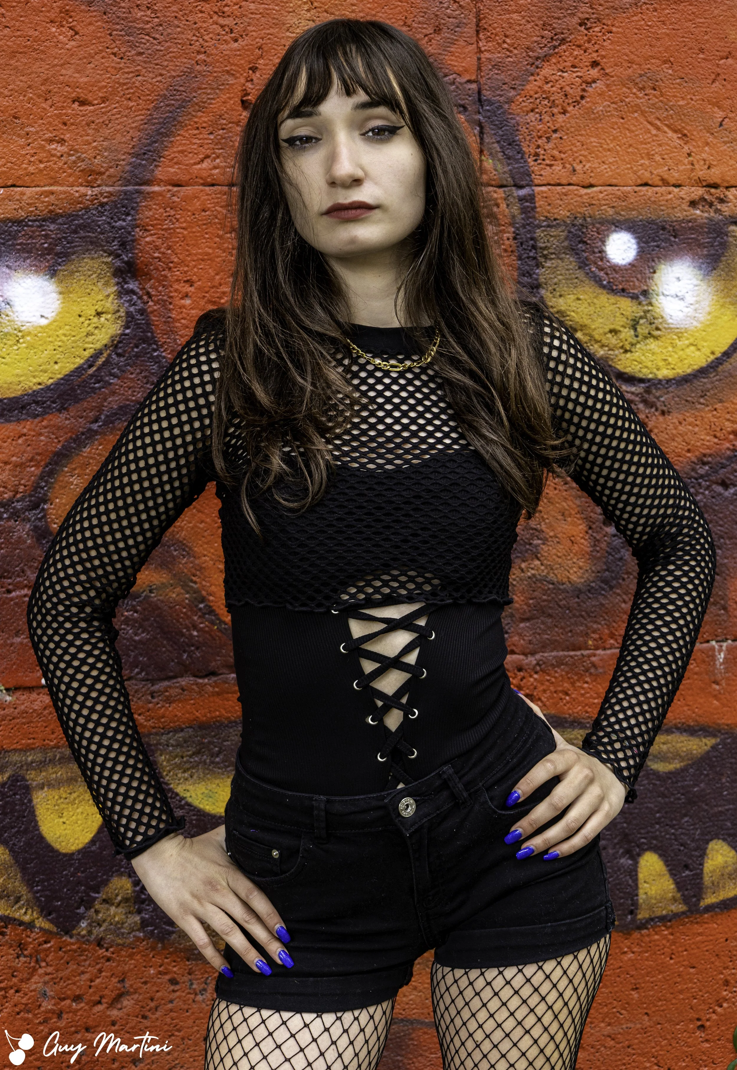 Ragazza con cardigan a rete e pantaloncini neri, posa con le mani ai fianchi davanti a un muro graffiti rosso e arancione.