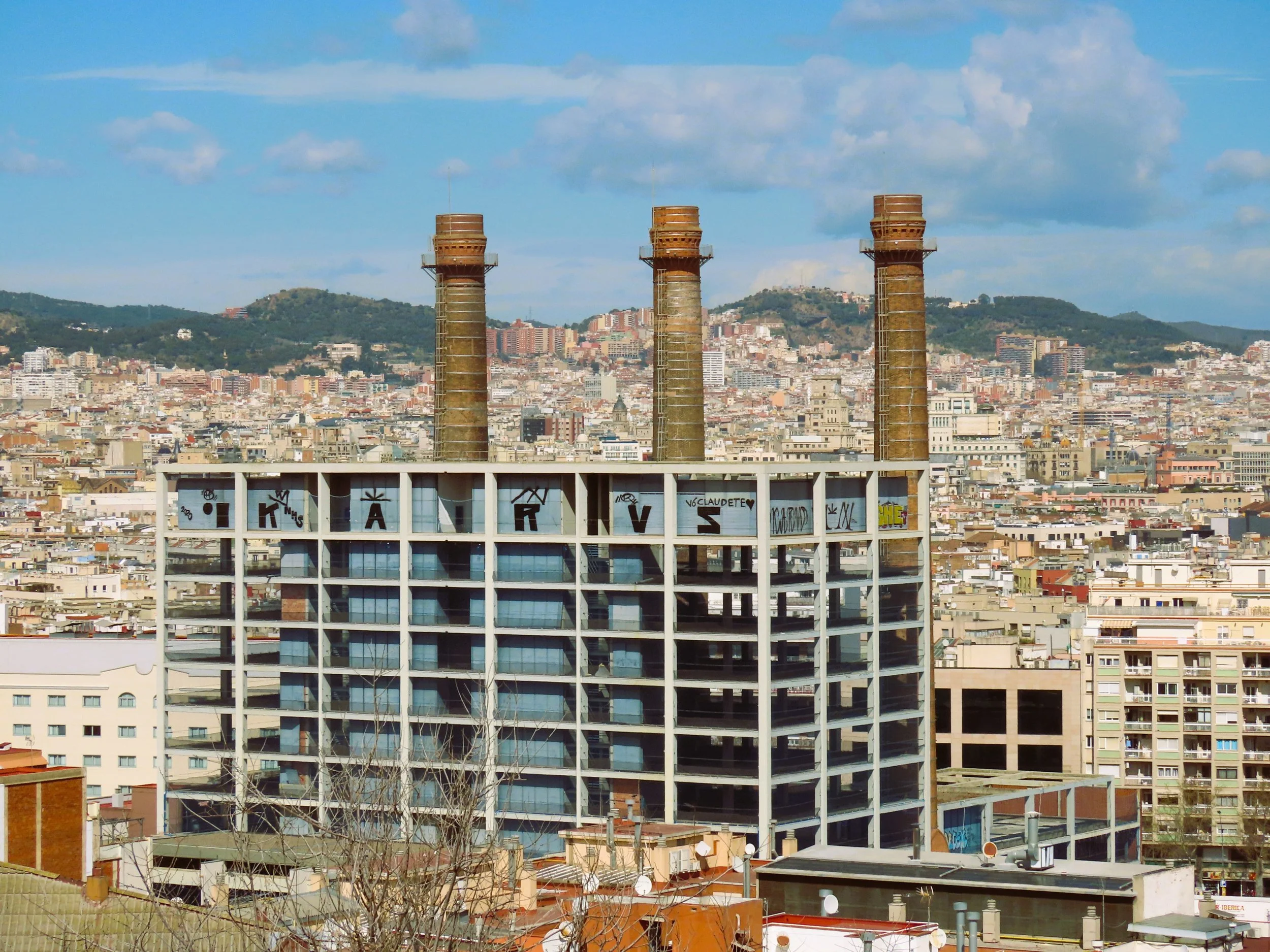 Edificio in costruzione con quattro grandi comignoli e graffiti sul tetto, con città urbana e colline sullo sfondo.