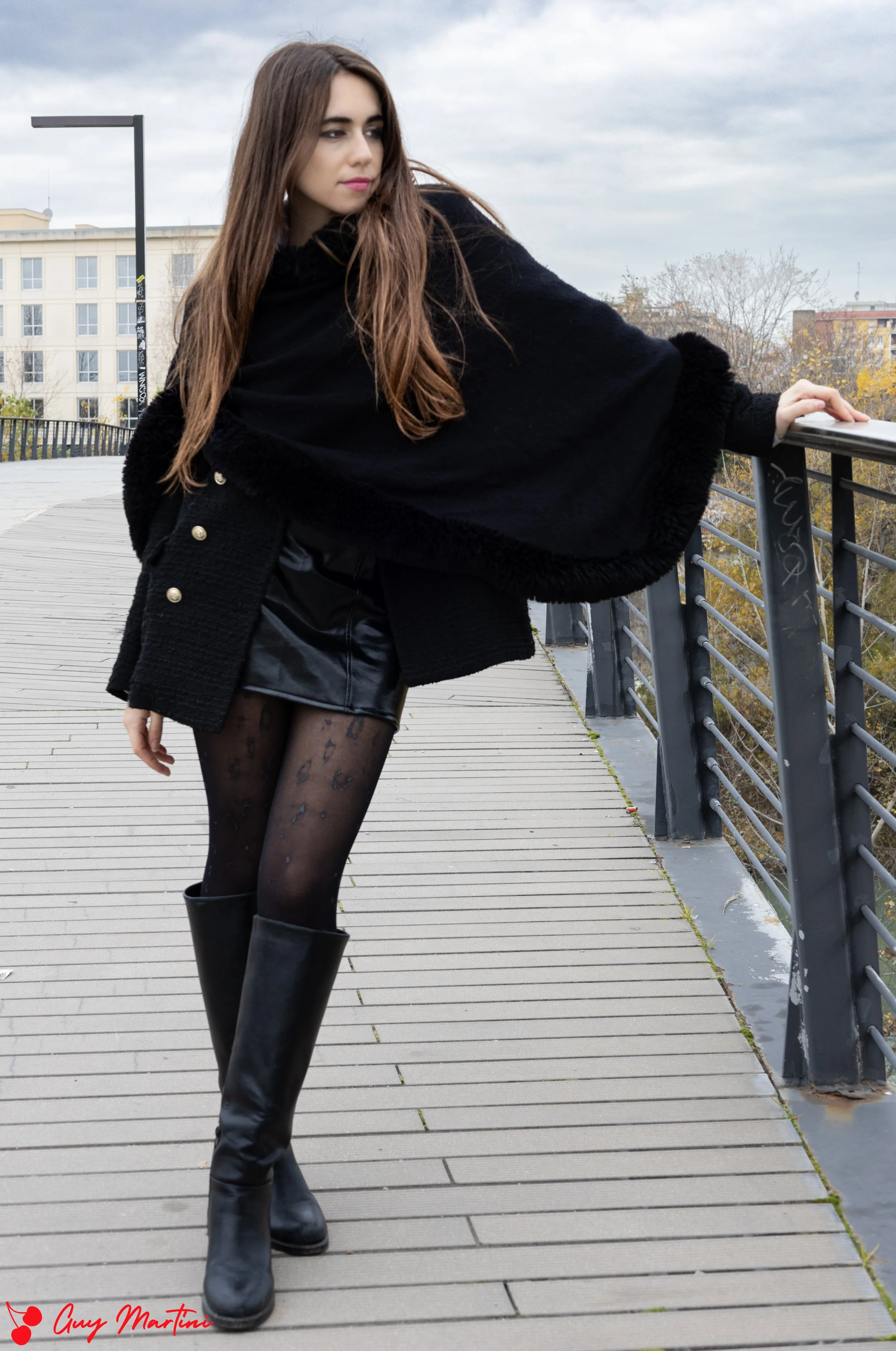 Donna con pantaloni neri, stivali alti e giacca nera, sulla passerella di un ponte, con un cielo nuvoloso dietro.