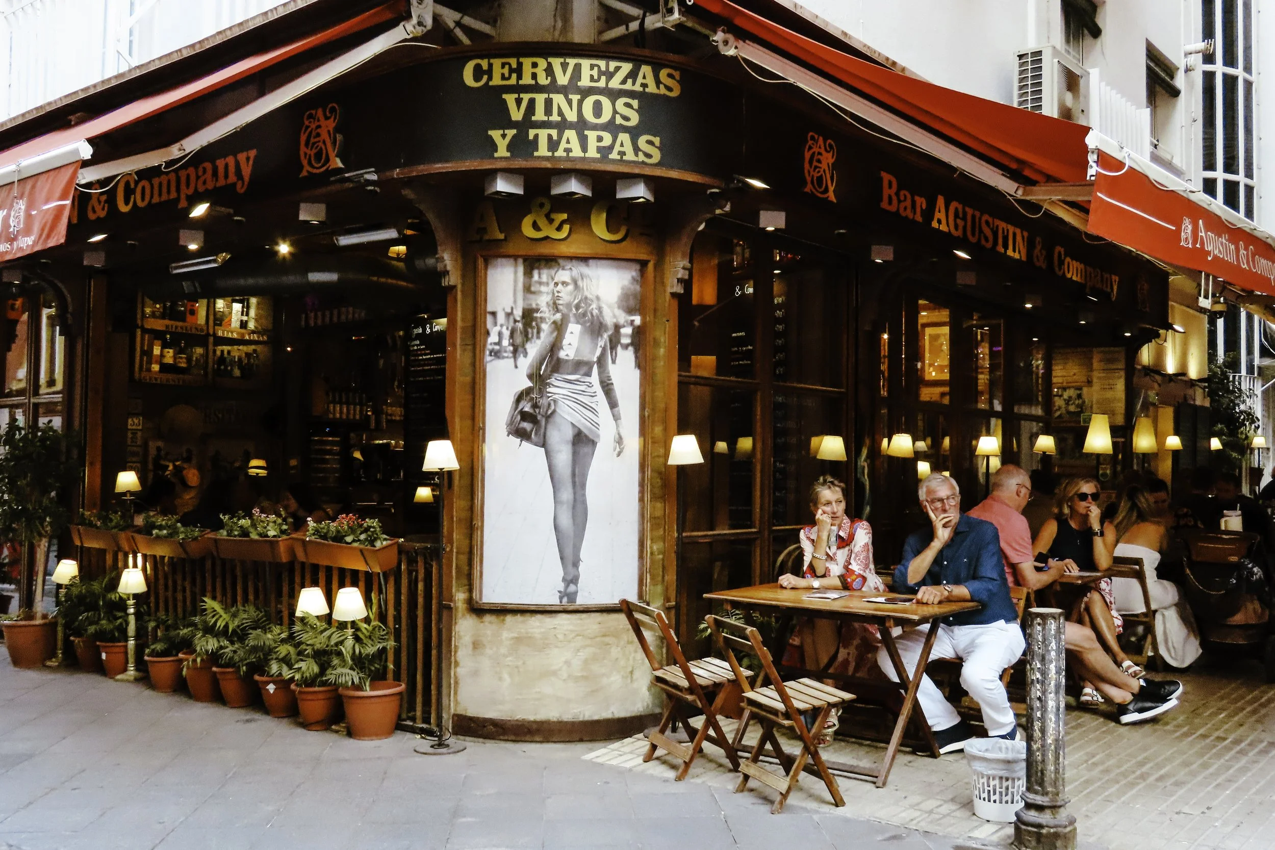 Esterno di un bar con tavolini e clienti seduti, piante in vaso e luci calde, insegna con parole in spagnolo e grande poster di una donna sulla parete